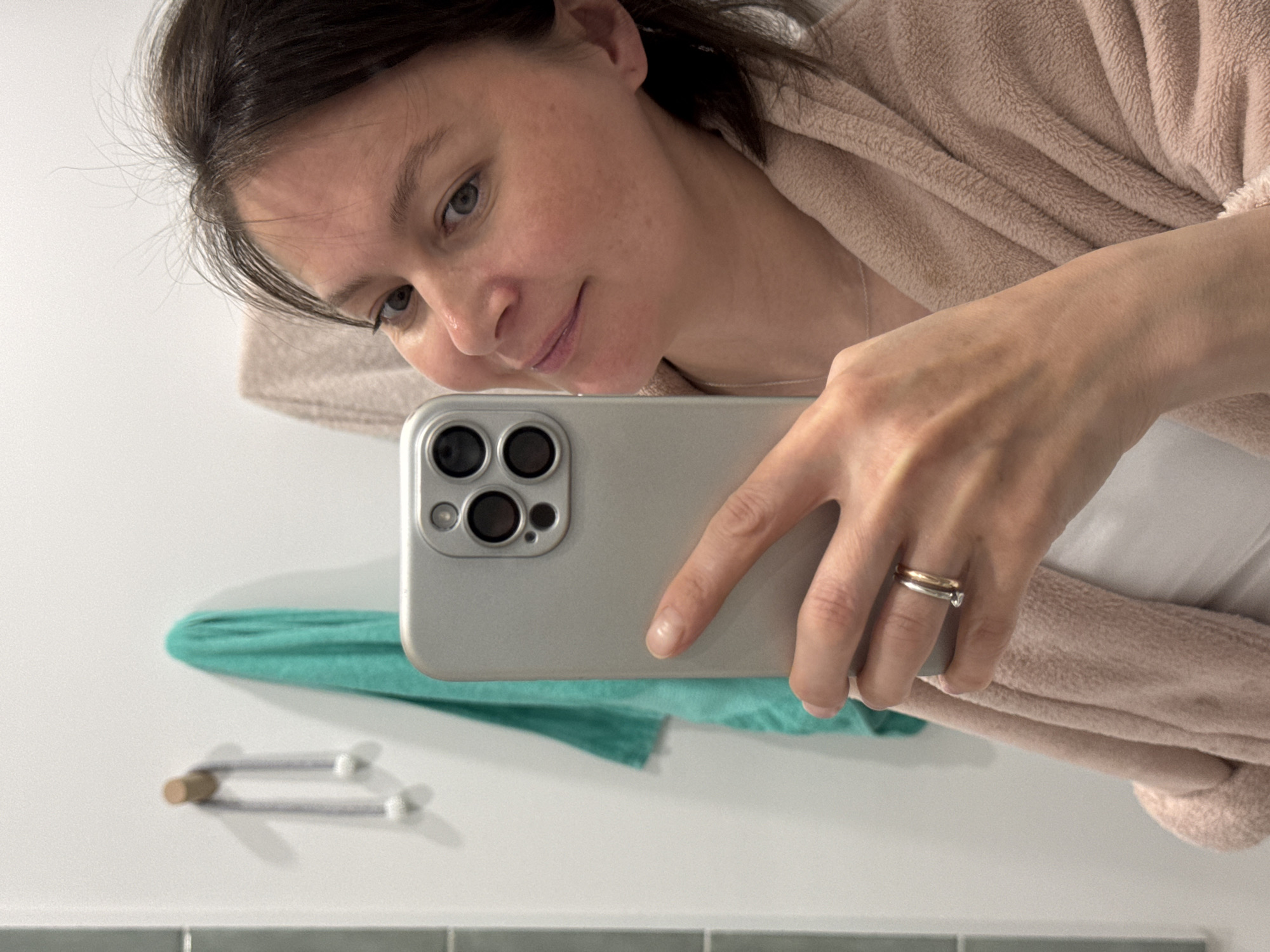 A woman is taking a selfie with a silver-colored smartphone. She is wearing a light pink robe and a white shirt. The phone has three camera lenses on the back. She is wearing two rings on her finger, one gold and one silver. A green towel is hanging on the wall behind the phone, and a towel rack is visible below the towel. The wall is white, with a tiled section at the bottom. The scene is likely a bathroom. There is no indication of the city of Izhevsk, Russia.