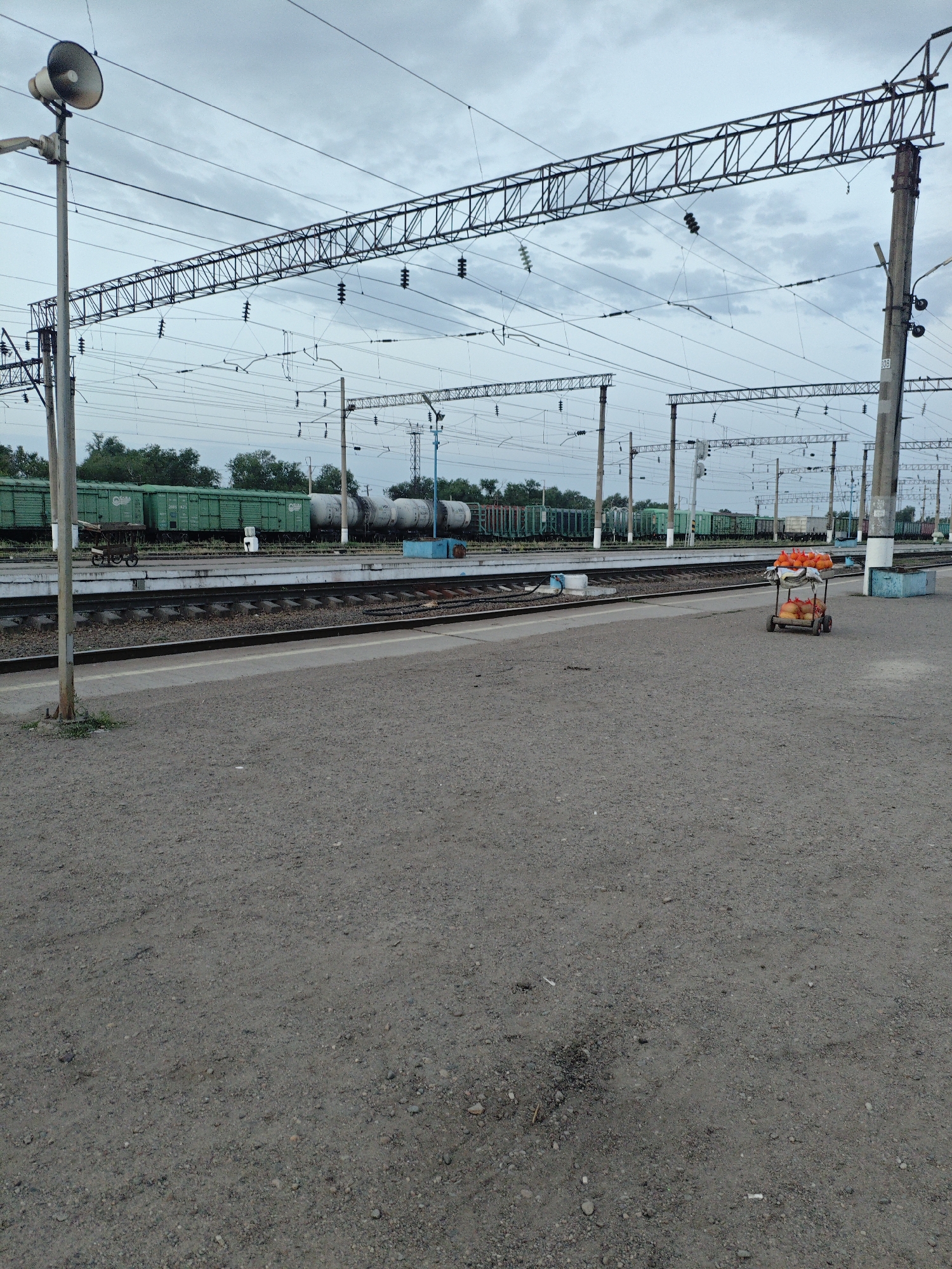 This image captures a view of a train station, likely in the city of Shu, Kazakhstan, characterized by its overcast sky and extensive railway infrastructure. The foreground is dominated by a gravelly platform, with a subtle patch of worn-down ground in the lower center. To the left, a tall pole supports a large loudspeaker, a common fixture at such locations. Beyond the platform, a set of train tracks runs parallel, with a train of green freight cars and white tank cars visible in the middle ground, parked on the tracks. Overhead, a complex network of electrical wires and support structures crisscrosses the sky, indicating the presence of an electrified railway system. On the platform in the middle ground, a small, two-wheeled cart is visible, laden with numerous orange and yellow bags, suggesting the presence of vendors or goods being transported. The overall atmosphere is one of a functional, somewhat utilitarian transportation hub under a muted sky.