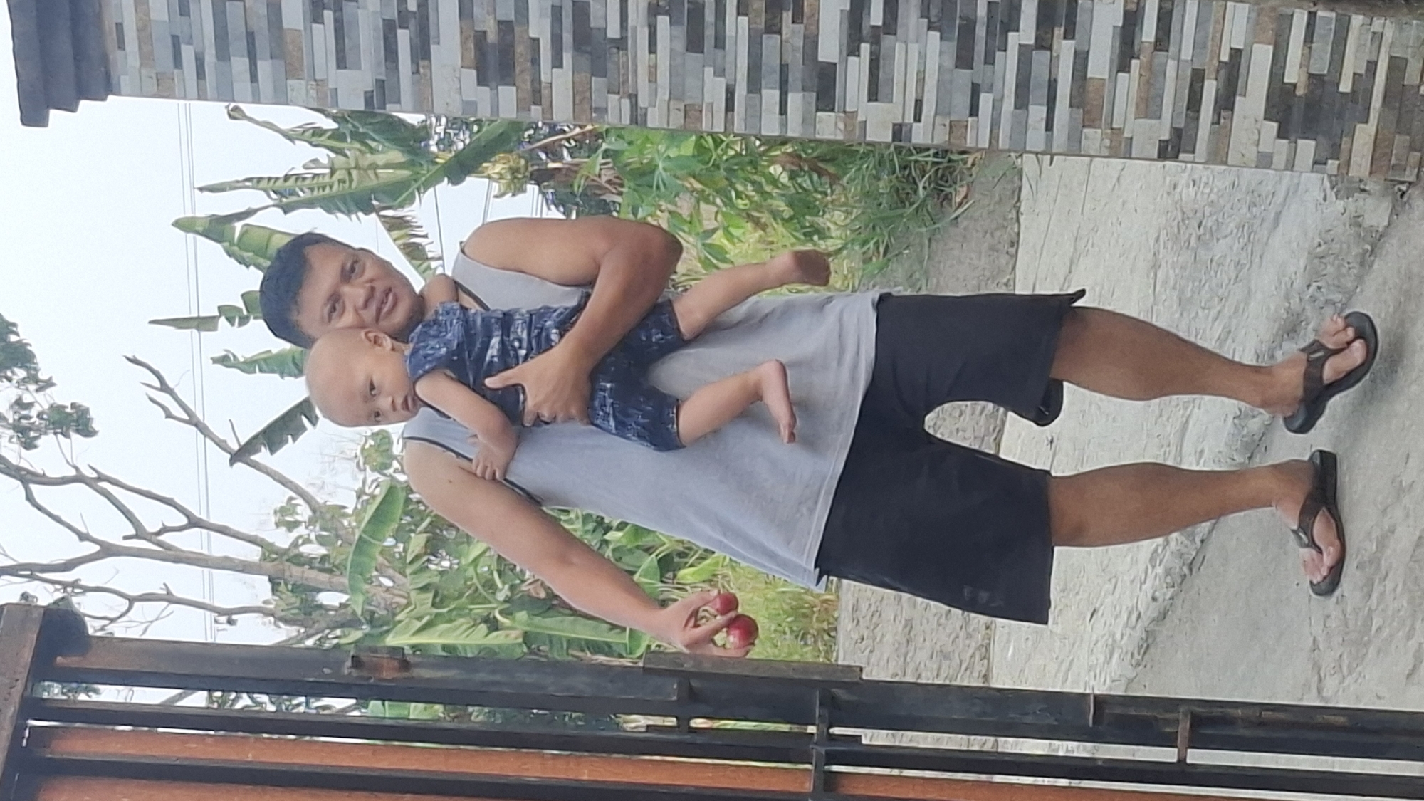 A man is carrying a baby on his left shoulder while standing next to a grey and brown brick wall. The man is wearing a grey tank top and black shorts. The baby is wearing a dark blue and white patterned outfit. In his right hand, the man is holding two red spherical fruits. The scene appears to be outdoors, with trees and foliage visible in the background. The ground is a light grey concrete or stone surface. The image is shot from a slightly low angle. The location is identified as Cibinong Hilir, Indonesia.