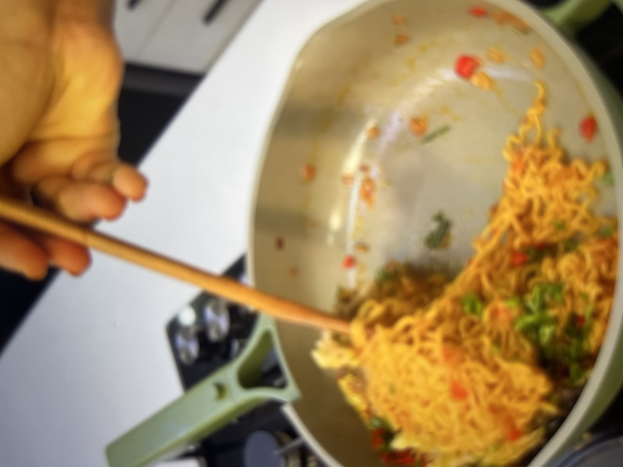 A close-up shot captures a hand holding a wooden chopstick, stirring a pan of noodles. The noodles are a vibrant orange, interspersed with green herbs and red chili flakes. The pan, a pale green, sits on a stovetop. In the background, a blurry kitchen counter and appliances are visible. The scene appears to be a home kitchen, likely in Abuja, Nigeria, as evidenced by the general aesthetic and common culinary practices associated with the region.