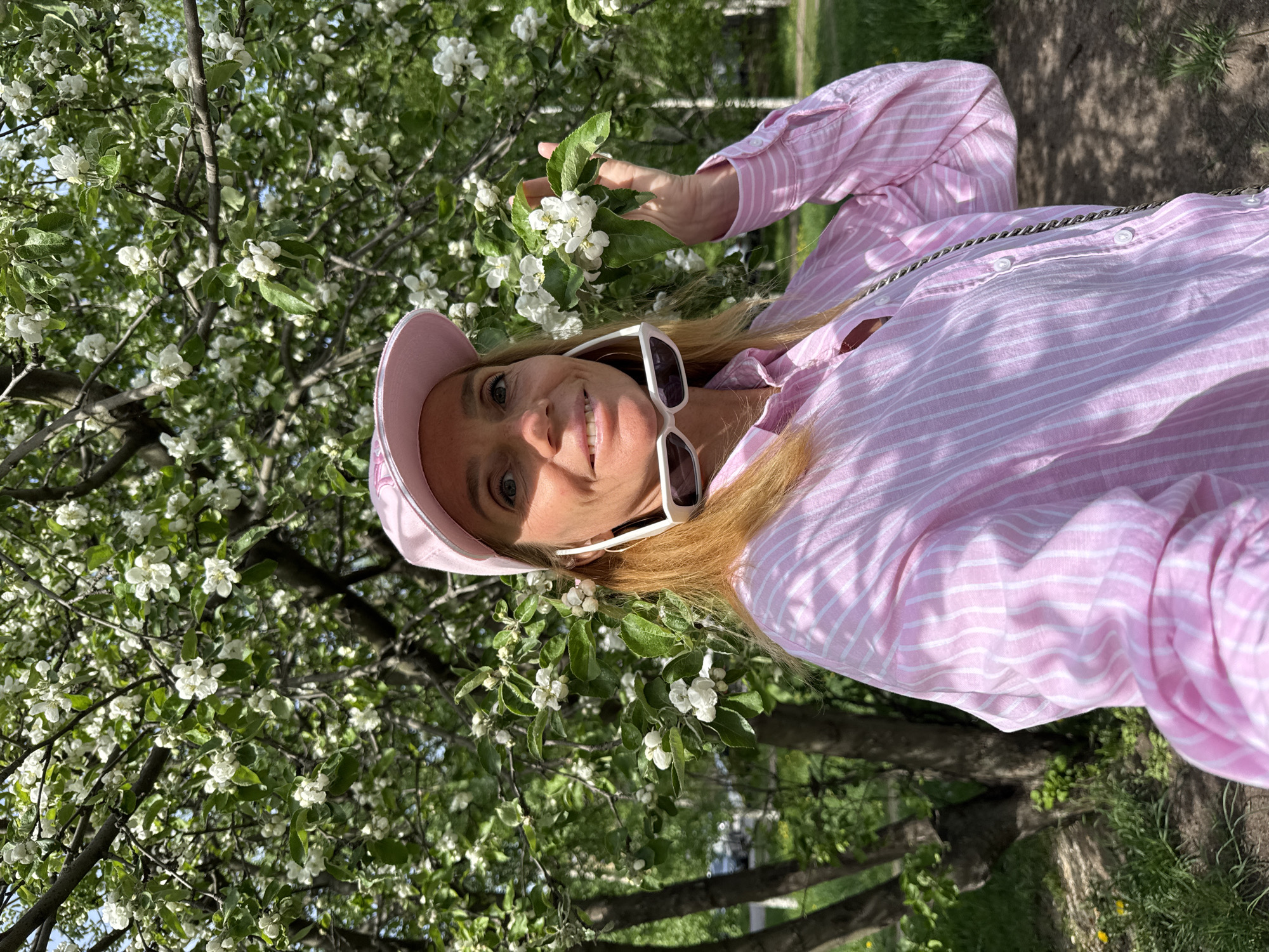 A woman stands by a flowering tree in Saint Petersburg, Russia. The woman wears a pink-and-white striped shirt, a pink baseball cap, and white-framed sunglasses. She is smiling and holding a branch of the flowering tree. The background includes other trees and foliage.