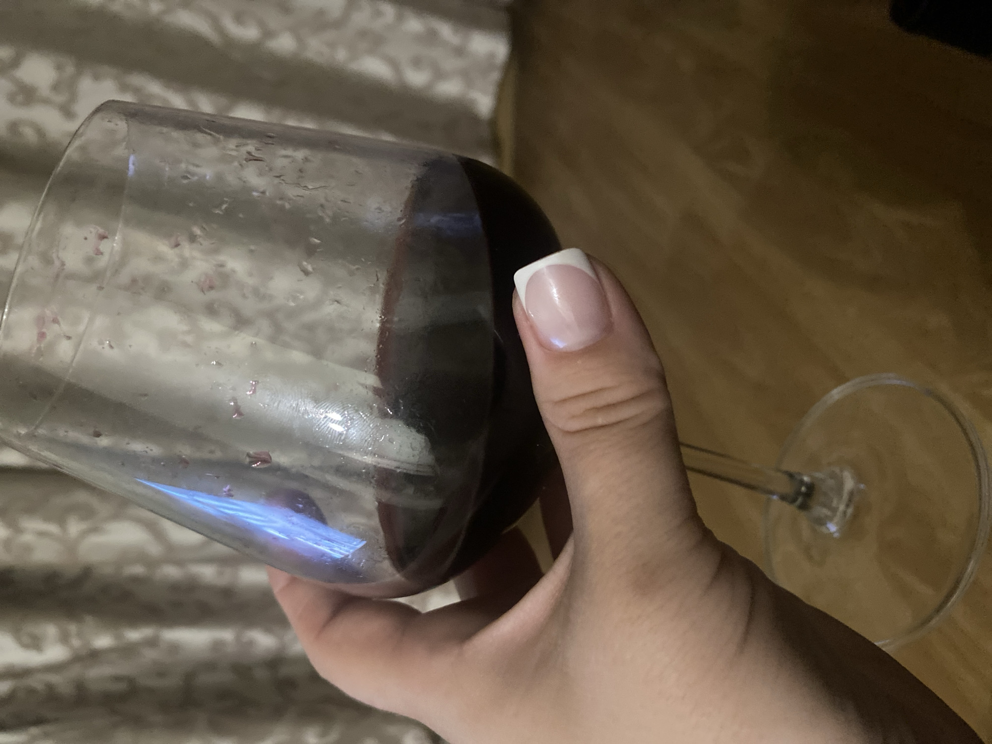 A hand holds a wine glass containing red wine. The glass has condensation and residue on the inside. The background includes a patterned fabric and a wooden surface. The location is not specified.
