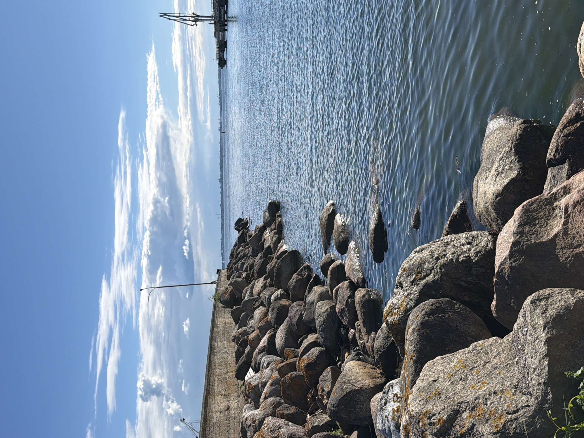 The image depicts a rocky shoreline next to a calm body of water, likely a sea or large bay, under a partly cloudy blue sky. In the foreground, a dense arrangement of large, weathered rocks forms the bank. Some of these rocks are partially submerged in the water. A concrete wall runs along the top of the embankment. Further out in the water, a pier with a tall crane is visible, along with a distant shoreline with industrial structures. A single lamppost stands on the wall near the left side of the image. The scene appears to be a coastal area, possibly a harbor or a section of the coastline in Viimsi, Estonia, known for its maritime character and rocky shores. There are no people or animals visible in the image. The overall impression is one of a quiet, natural waterfront with hints of industrial activity in the distance.