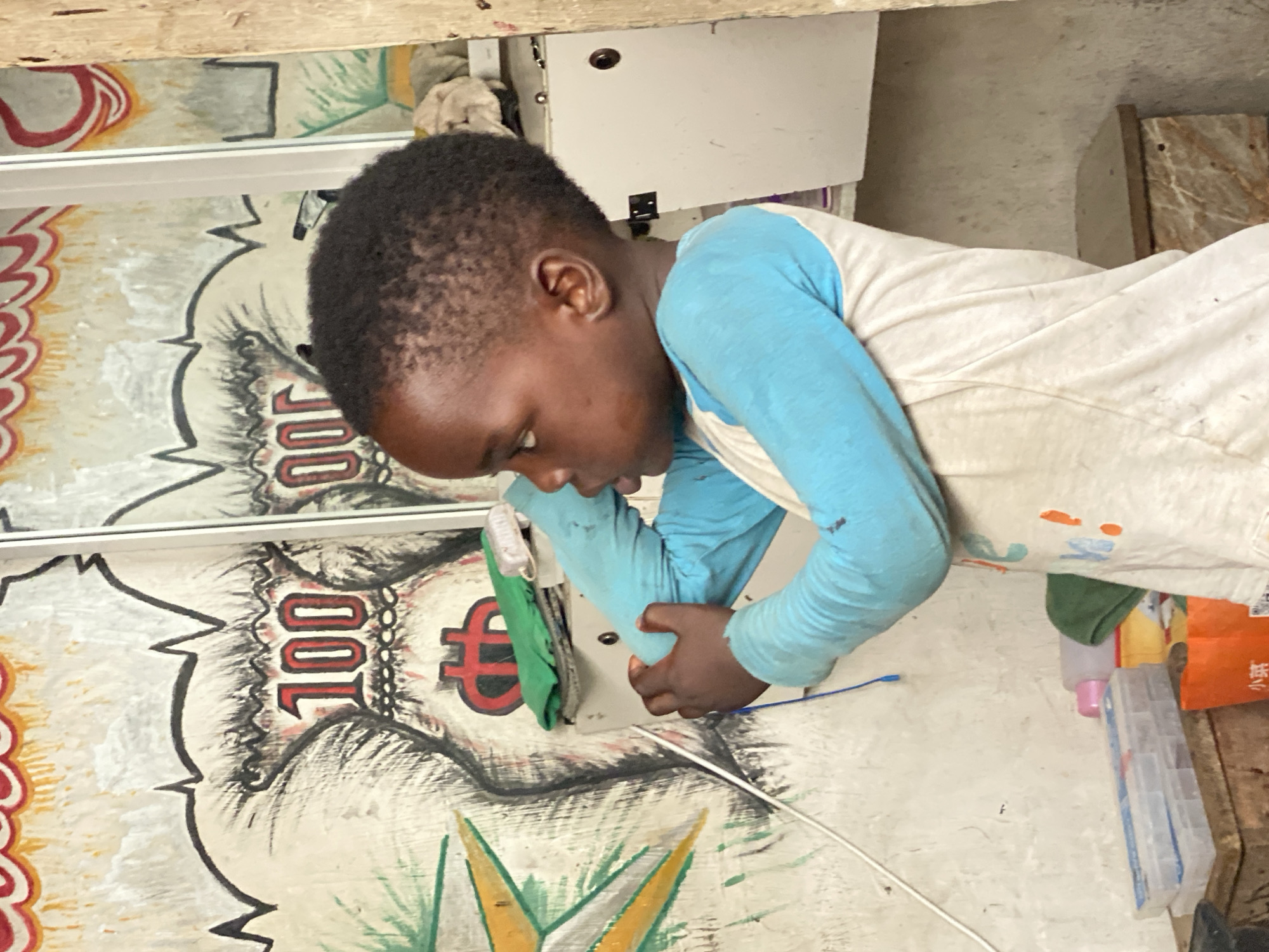 This image, taken in Lekki, Nigeria, features a young boy intently focused on an object or task. The boy, with short dark hair and dark skin, is wearing a long-sleeved shirt with a white torso and light blue sleeves. He is leaning forward, with his hands positioned on or around a white piece of equipment or a machine, which has a dark circular element and some metallic components. A green fabric is visible near his hands.
The background consists of a painted surface adorned with stylized currency motifs, including the number "100" and a symbol resembling a dollar or Euro sign. These designs are repeated and reflected in a narrow, horizontal strip of glass or mirror located above the main painted area. The overall setting appears to be an indoor, possibly commercial or workshop environment, characterized by a functional and somewhat rustic aesthetic.
To the right, partially visible objects include a pink bottle, a clear plastic container with dividers, and an orange package, alongside various other miscellaneous items. A white wire extends across the lower part of the frame, and a wooden beam is visible at the top. The boy's posture and gaze convey deep concentration on his activity, the specific nature of which is not entirely clear from the image. The time of day appears to be daytime, with indoor lighting.