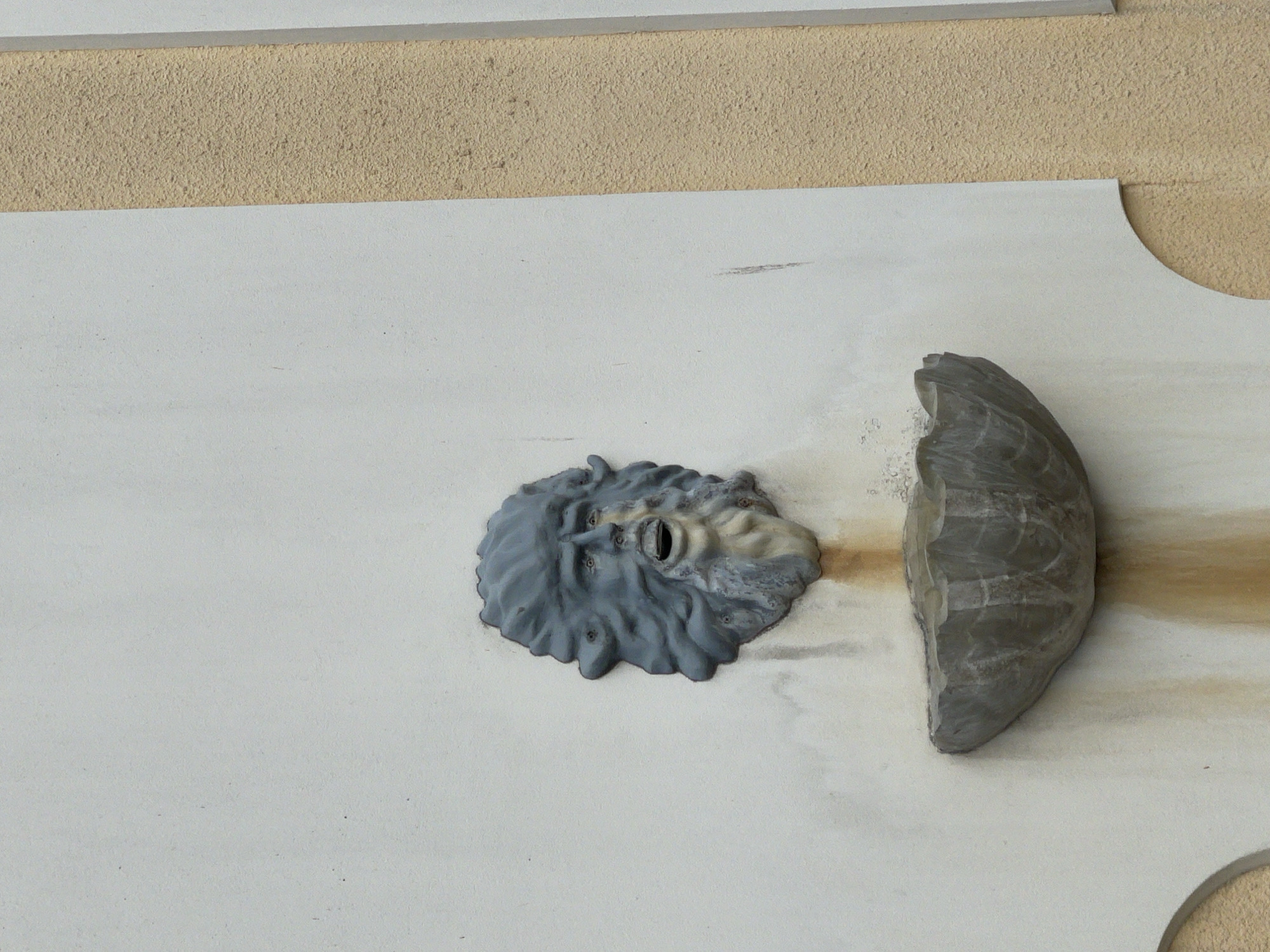 A grey, weathered fountain sculpted in the form of a face is mounted on a beige stucco wall. The face has a stern expression, with its mouth open as if to spout water. Below the face is a grey, shell-shaped basin designed to catch the water. Brownish streaks are visible on the wall below the basin, indicating water stains from past use. A darker, textured vertical element runs along the right side of the image, bordering the beige stucco. The overall scene suggests an outdoor setting, possibly a historical or decorative architectural element.