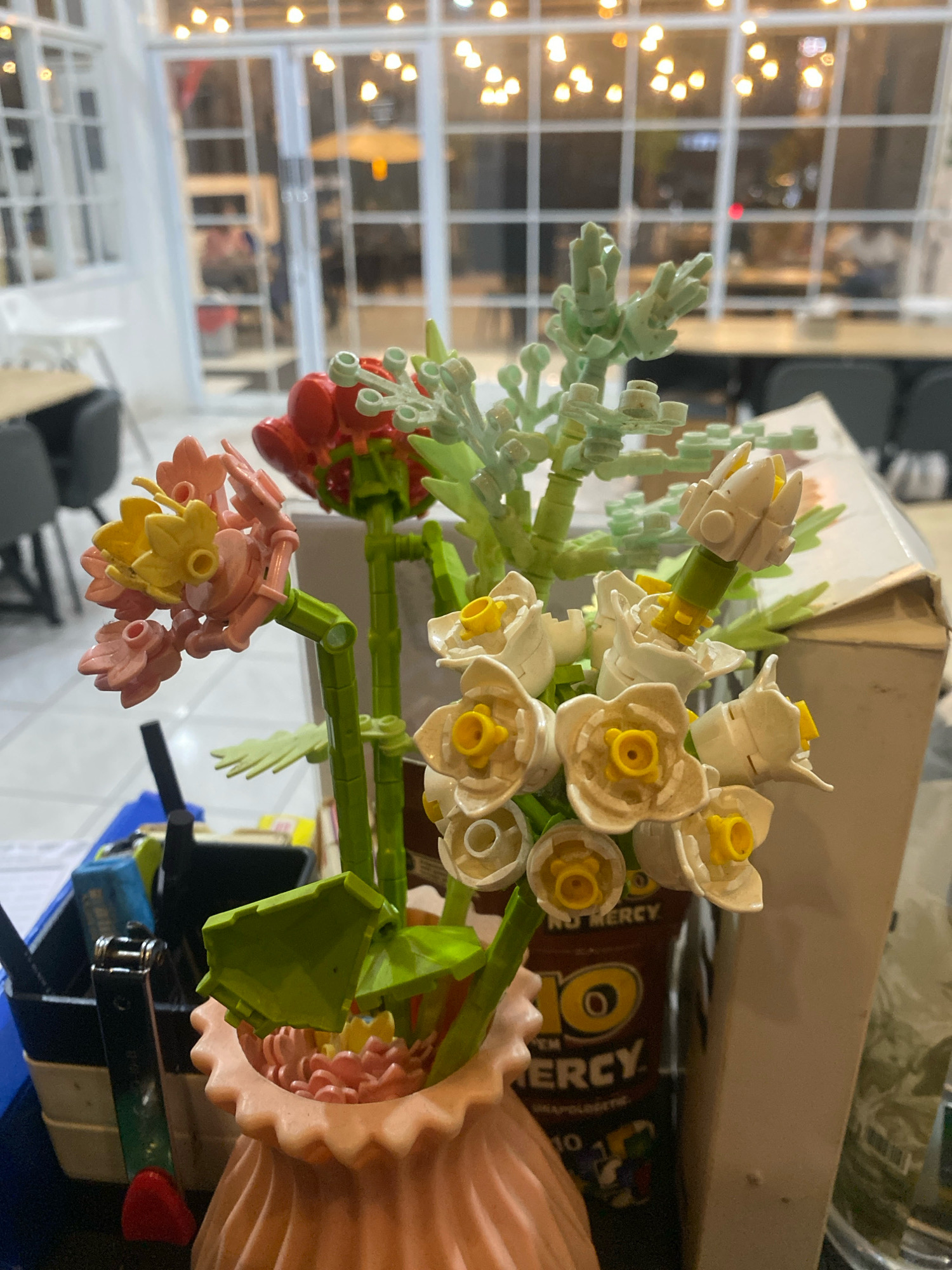 An indoor shot at eye-level captures a Lego flower arrangement on a table. The bouquet features white and yellow daffodils, pink and yellow flowers, and a red rose. The arrangement sits in a ribbed, pink vase. In the background, through glass doors with white grilles, a restaurant interior is visible with tables, chairs, and ceiling lights. A cardboard box with "NO MERCY" printed on it is to the right of the vase. A stapler and pens in a holder are visible on the left side of the table. The scene is well-lit, suggesting artificial lighting. The location is Sangatta, Indonesia.