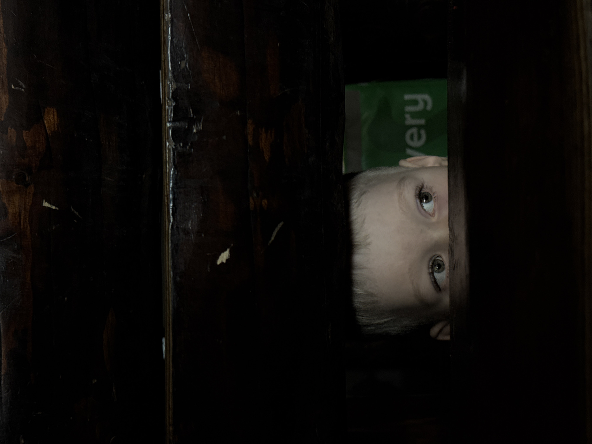 A close-up, low-angle shot shows a child's face peeking through a narrow vertical gap, presumably between two dark wooden panels. The child has light skin, blond hair, and blue eyes, and appears to be looking up and out of the space. A green object with the word "very" in white is partially visible behind the child's head. The surrounding environment is very dark, with the wooden panels dominating the scene. The location is not determinable from the image.