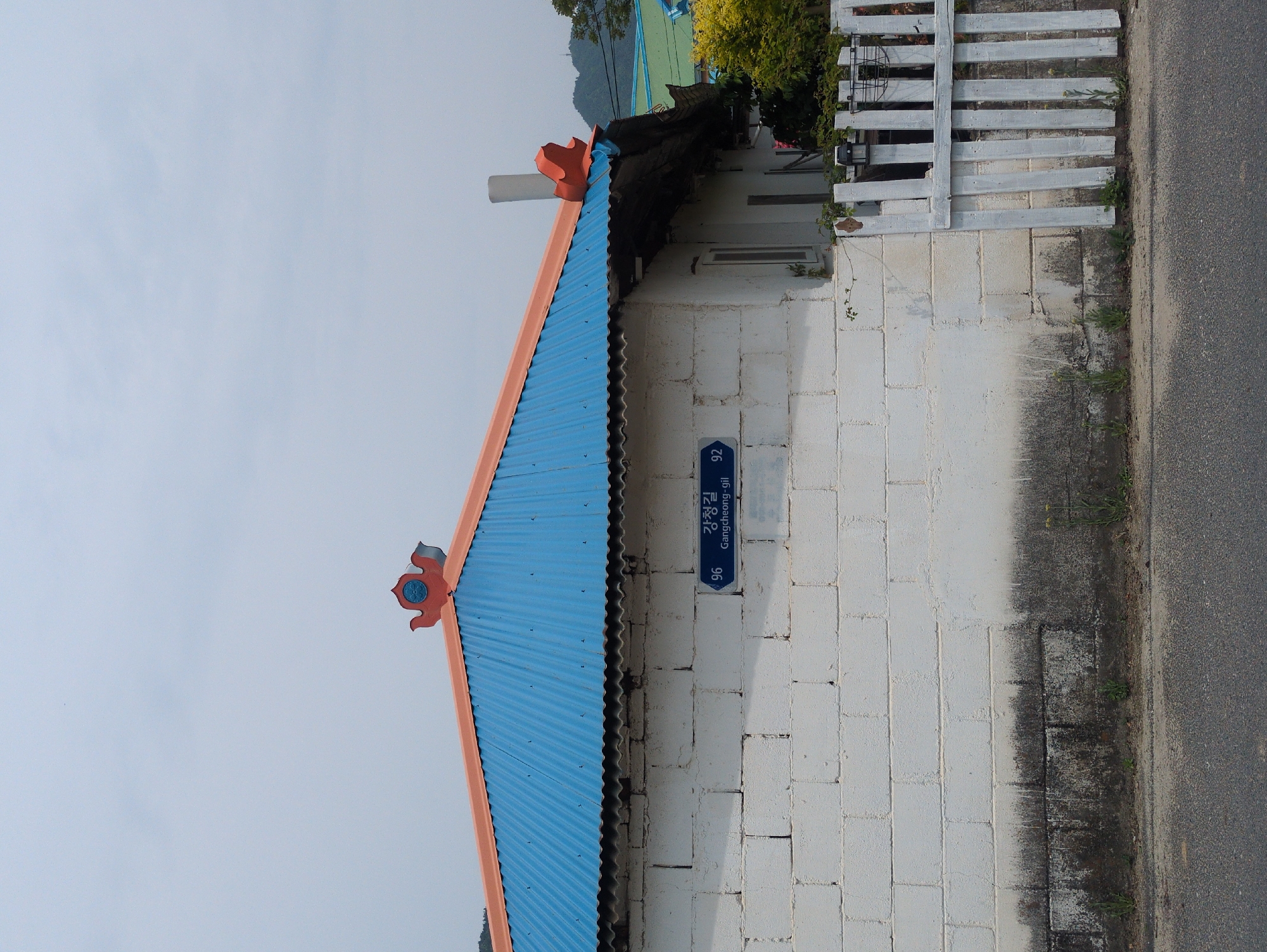 A medium shot depicts the side of a white brick building with a traditional Korean roof in Gangcheong-ri, South Korea. The roof is a blue corrugated metal with orange trim and decorative elements at the corners. A street sign reading "Gangcheong-gil" with the numbers 92 and 96 is affixed to the wall. The building is partially obscured by a decaying black roof overhang, and a white picket fence extends to the right. The foreground is a paved road. The sky is pale blue.