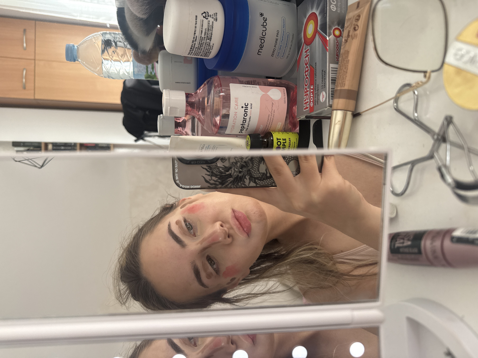 A woman is taking a selfie in a mirror. Her face is decorated with makeup. On the counter next to the mirror are various skincare and makeup products, including a bottle of water, Medicube Zero Pore Pads, Peptaronic toner, Nurofen Forte, and a tube of foundation. The background includes wooden cabinets. There is no indication that the image was taken in Blagoevgrad, Bulgaria.
