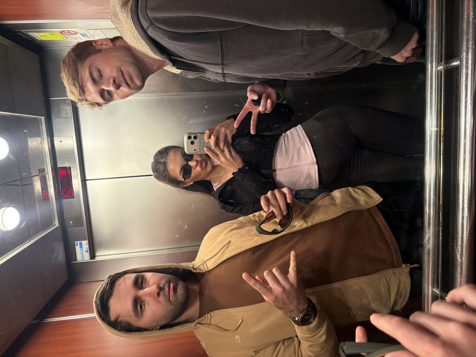 Three individuals are captured in a mirrored selfie taken inside an elevator.
The elevator interior features dark wood-paneled side walls and a reflective stainless steel rear wall that serves as the mirror. Two circular ceiling lights illuminate the space. Above the mirror on the rear wall, a red digital display indicates "5," likely representing the current floor. Below this, blue and white stickers are present, one appearing to be a "no smoking" sign. A chrome handrail is reflected horizontally across the bottom of the frame. The specific location within the city of Tel-Aviv, Israel, is not discernible from the visual evidence.
From left to right:
1. An adult male with dark hair and a beard, wearing a tan hooded jacket with subtle branding on the left chest over a brown t-shirt, and a dark watch on his left wrist. He holds dark-framed sunglasses in his right hand and performs a "rock on" hand gesture with his left hand. His expression is neutral.
2. An adult female with dark hair, wearing rectangular dark sunglasses, a black cropped jacket over a light pink structured top, and dark leggings. She is taking the photograph using a light-colored smartphone with multiple camera lenses, held in her right hand, while pouting her lips towards the camera.
3. An adult male with light-colored hair, wearing a dark gray hooded jacket with a cream-colored sherpa lining. He performs a "peace" hand gesture with his left hand. His expression is a slight smirk.