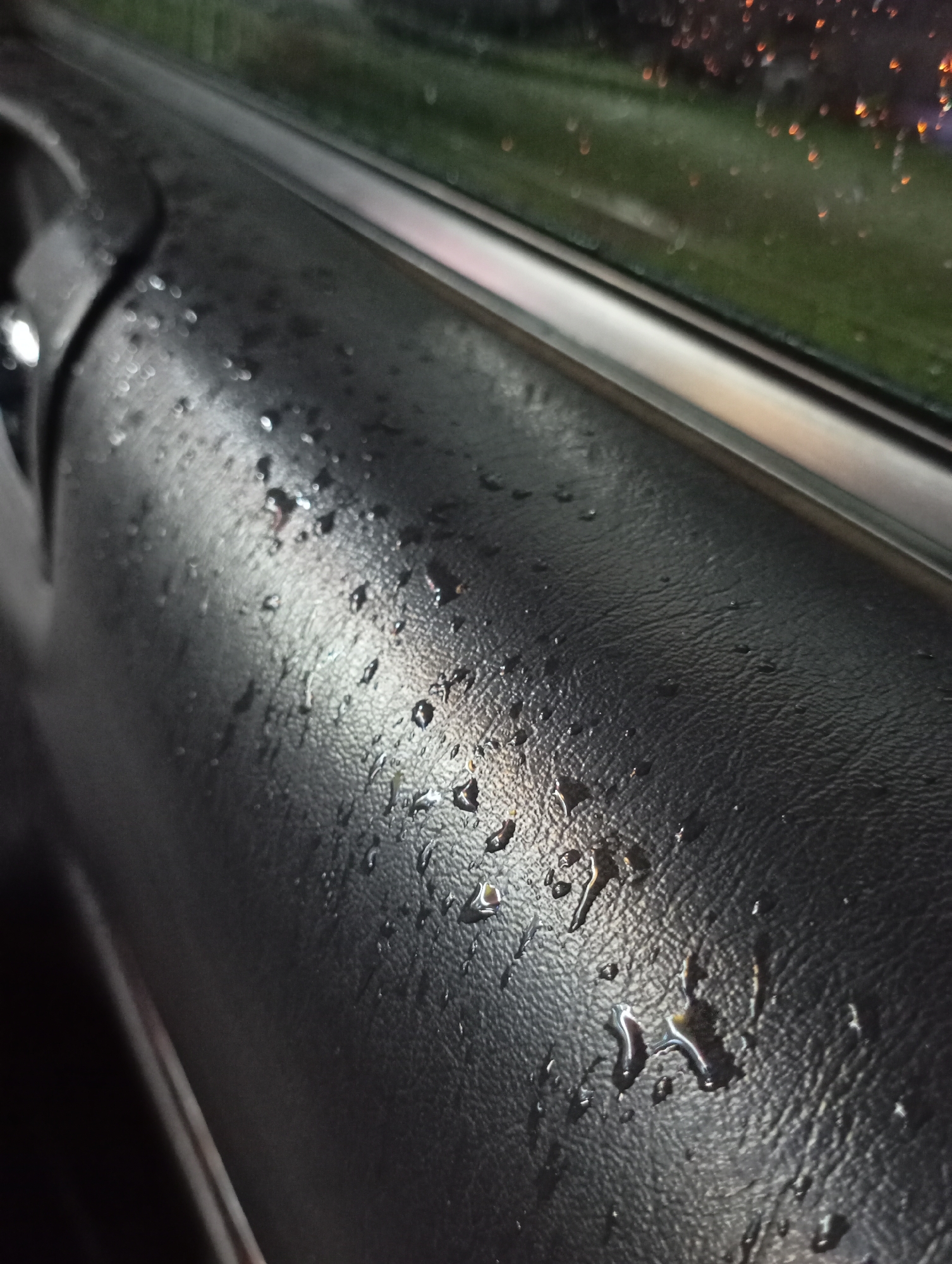 The image shows the interior of a car, specifically a black leather or vinyl-covered portion of a car door. The surface is wet, with droplets of water scattered across it. The window is visible in the upper part of the image, with blurred lights outside, possibly from street lamps or other vehicles. The scene appears to be taken at night or during a low-light situation, as the interior is dark. There is no indication of location, including Industrialny, Russia, or people within the image.