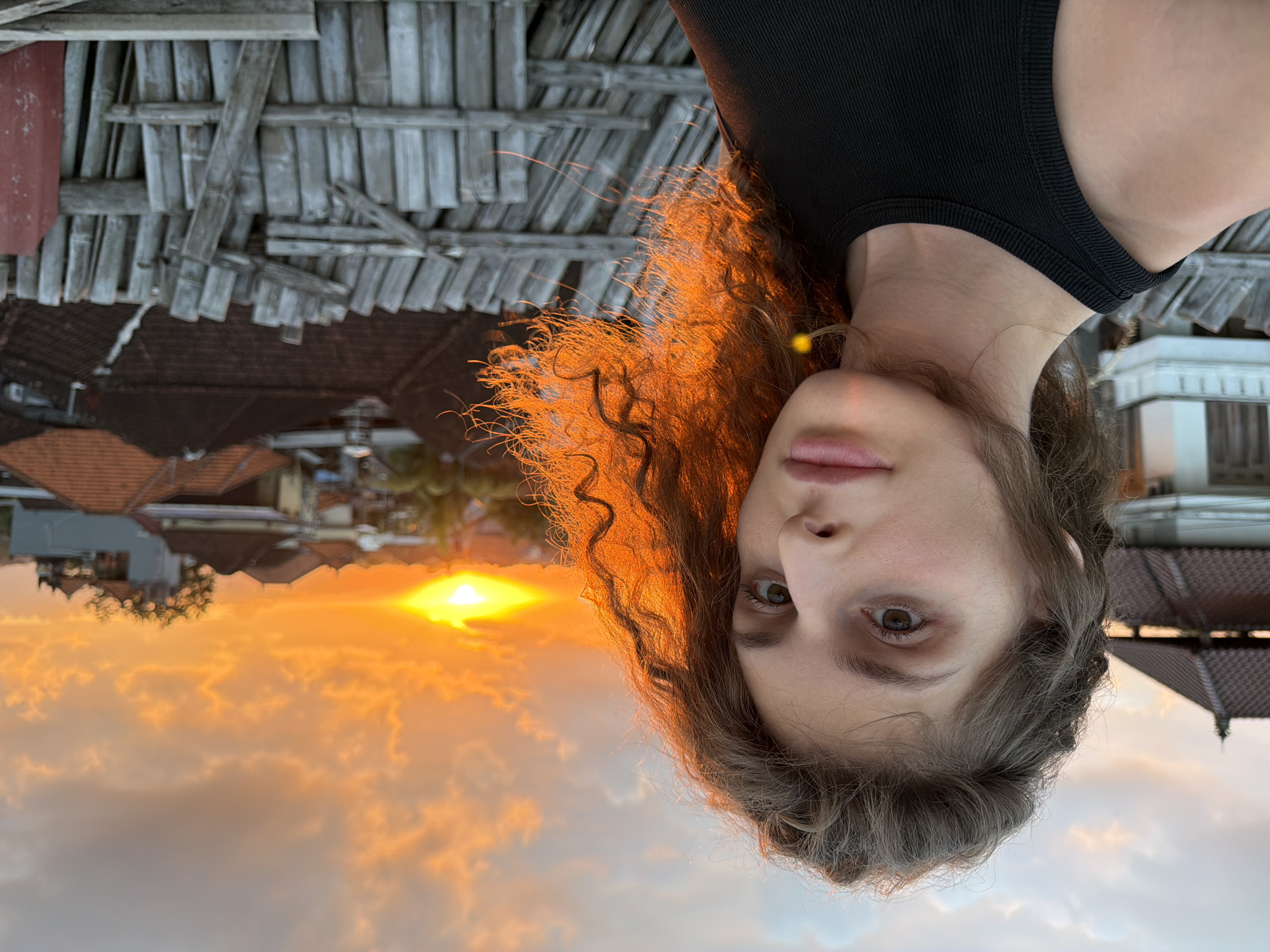 An outdoor, close-up, right-facing selfie of a young woman with curly, brown hair is captured against a vibrant sunset sky. She is wearing a black tank top. The background features a cityscape with buildings and rooftops, illuminated by the warm orange glow of the setting sun. A wooden fence is visible in the foreground. The location is North Kuta, Indonesia.