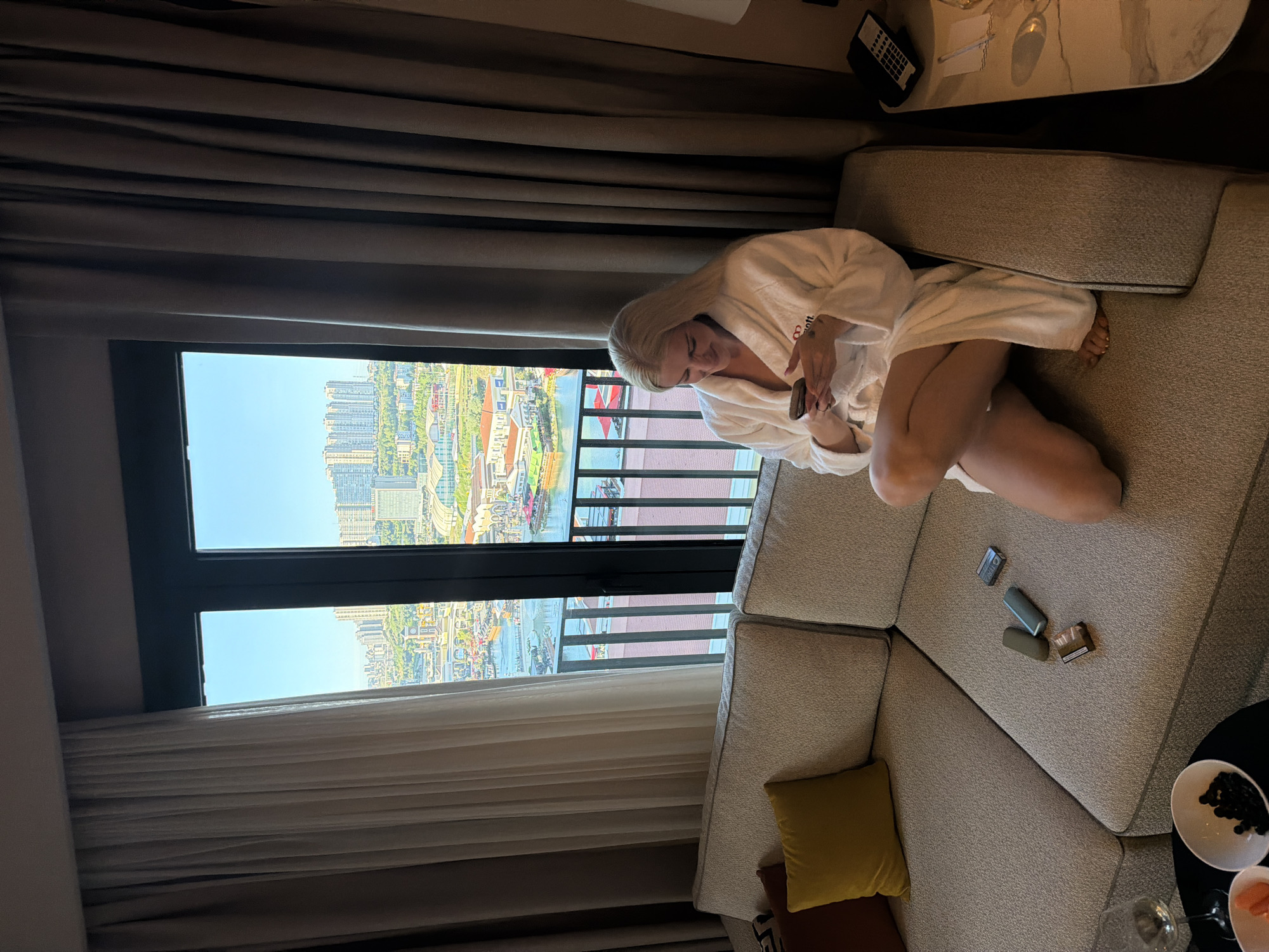 A blonde woman in a white robe sits on a gray sectional sofa in a hotel room with a view of Küçükçekmece, Turkey. The woman holds a small object in her hands and looks down. On the sofa near her are several small items, including what appear to be vape cartridges and a pack of cigarettes. A yellow decorative pillow and the corner of a small table with two plates and glasses are also visible. The room's window offers a view of a cityscape with buildings and a body of water. The view is from high up, presumably from a hotel room.