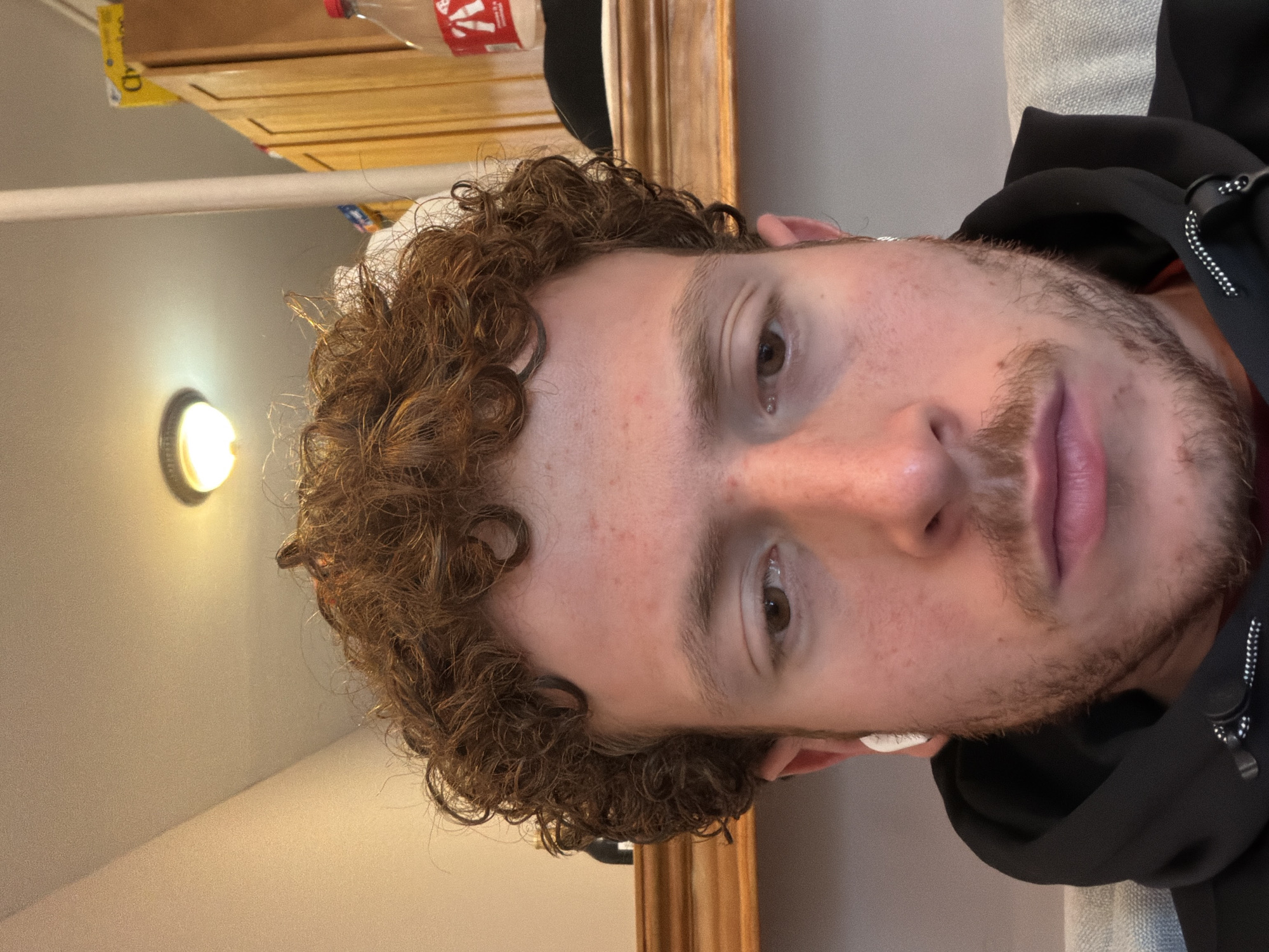 A close-up portrait features a young man with light skin, brown curly hair, and brown eyes. He has a light beard/stubble on his chin and jawline, along with scattered freckles on his forehead and cheeks. He is wearing white earbuds and a dark hooded top. His expression is neutral as he gazes directly towards the viewer.
The setting is indoors, with a warm, artificial light source illuminating the scene from the upper left. Behind the man's head and to his left, a wooden cabinet or shelf is visible. On this surface, a red bottle, consistent in color and shape with a Coca-Cola product, is present, along with a yellow item that appears to be packaging, partially revealing the letter 'D'. To the right of the man's head, a section of an ornate, golden-toned wooden frame, possibly from a mirror or picture, is visible. Behind his right shoulder, a textured, light-colored fabric, such as upholstery, can be seen. The ambient lighting suggests a cozy indoor environment.