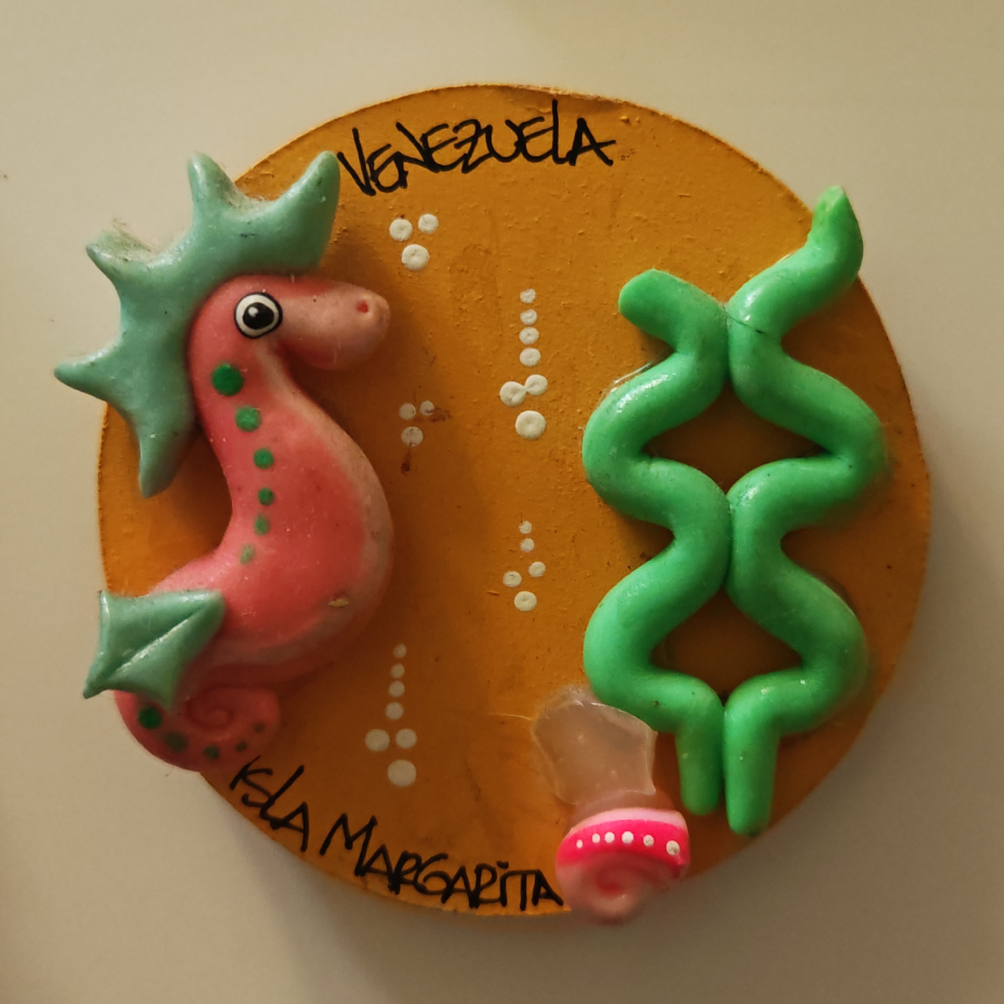The image displays a close-up of a souvenir, likely a refrigerator magnet, crafted on an orange, circular base with a textured finish. The central subjects are a pink seahorse figure and a wavy, bright green, plant-like form, possibly representing seaweed or coral.
The seahorse, positioned on the left, is predominantly pink with light green fins and dorsal spikes, and small green dots on its body. It features a distinct black and white eye. To the right, the green plant-like element twists upwards, forming an hourglass shape. Scattered white dots across the orange background suggest bubbles. A small, pink and white striped cylindrical piece, possibly part of another decorative marine element, is adhered to the bottom right of the base with visible clear glue. Both the seahorse and the green plant show minor accumulations of dust.
The souvenir features black handwritten text on its surface: "VENEZUELA" is inscribed across the top, and "ISLA MARGARITA" is written along the bottom curve, indicating the object's origin or theme is related to Isla Margarita, an island in Venezuela. No activity is taking place; the image is a still life of this decorative item. There are no visible cues for time of day or weather.