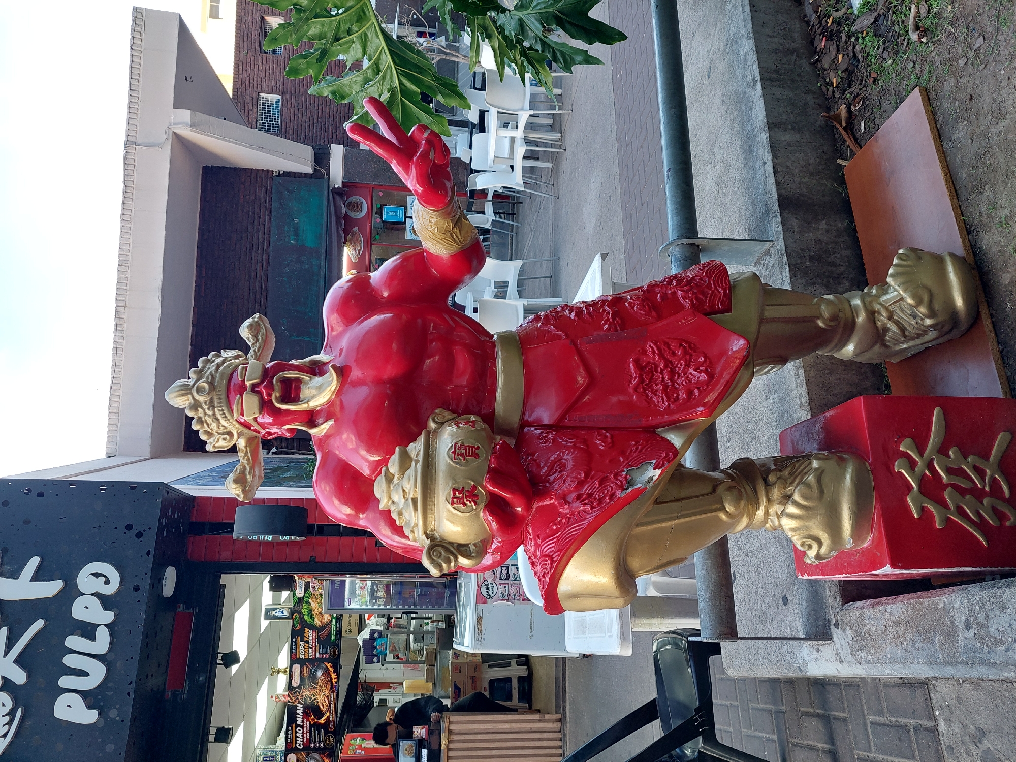 This media file presents an outdoor scene in Buenos Aires, Argentina, featuring a large, ornate red and gold statue prominently displayed outside what appears to be an Asian restaurant. The statue depicts a muscular figure, possibly a Chinese deity or guardian, adorned in traditional-style armor and a golden headpiece. Its right hand is raised, forming a V-sign, and its chest and base are decorated with gold Chinese characters, including "聚寶" (jù bǎo - gathering treasures) and "發" (fā - prosper/wealth).
The setting is a commercial area, brightly lit, indicating daytime. To the left of the statue is a black storefront sign with "PULPO" in white text. Through the entrance, menu boards are visible inside, clearly displaying "CHAO MIAN" and "SOPA WAN TAN CON FIDEOS", confirming the Asian culinary theme. A person can be seen working behind a counter within the restaurant.
On the right side of the image, an outdoor seating area with multiple white tables and chairs extends into the background. Overhead, green foliage from a plant or tree adds a touch of nature to the urban environment. The ground is a mix of paved sidewalk and dirt patches. Additional text visible on various signs or objects includes "INI", "ColonoPlas", "BULDO", and "PILI DO", likely brand names or labels on refrigeration units. The overall impression is a vibrant street-side eatery.