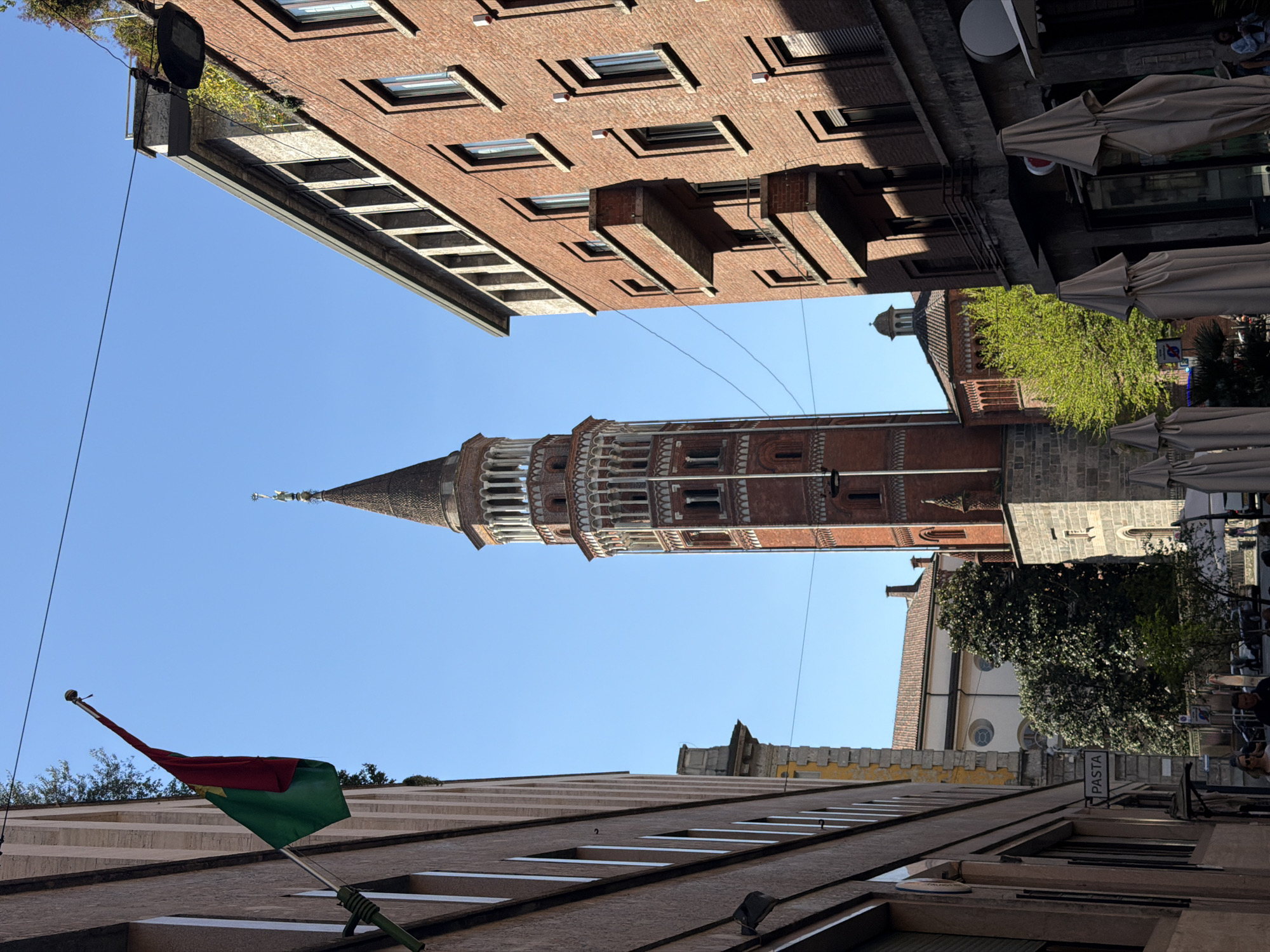 An eye-level, rotated, long shot captures a view of Milan, Italy, looking upwards from a narrow street. The Torre Velasca tower is centrally positioned in the shot, towering above the surrounding structures with its distinctive architectural style. The sky is a clear, bright blue, contrasting with the reddish-brown brick of the tower. Two multi-story buildings with numerous rectangular windows line the top and bottom of the view, framing the tower. A flag, with a dual-colored design, green and red, is affixed to the building at the bottom. On the right side of the scene, tan-colored umbrellas are visible, suggesting an outdoor dining area. A tower-like structure sits to the right of the Velasca tower. A sign marked "PASTA" can be seen in the lower right.