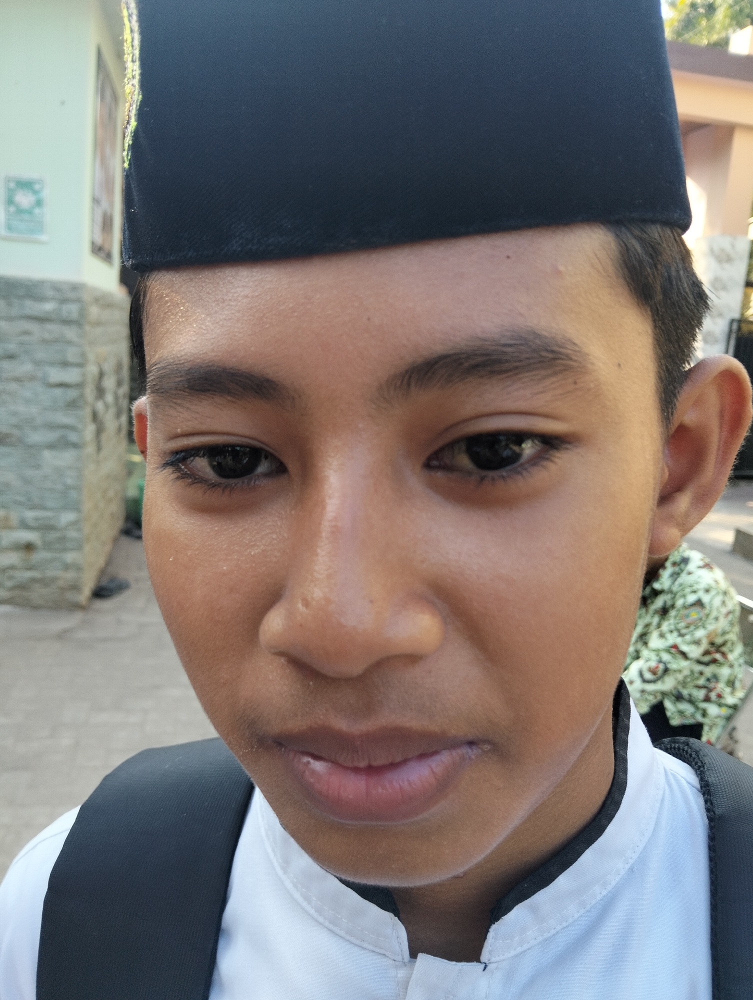 The media file is a close-up, eye-level, medium shot of a young person wearing a black peci hat. The subject is facing the camera, with their head tilted slightly. They have dark eyes, dark eyebrows, and dark hair visible around the edges of the peci. Their skin is a warm brown tone, and they appear to have a slight sheen, possibly from sweat. The subject is wearing a white shirt with a black collar, and a black backpack is visible over their shoulders.
In the background, to the left, there is a light-colored building with a stone wall and a sign with green and white graphics. Further back, to the right, is another building with a peach-colored wall and some greenery. The lighting suggests it is daytime, likely outdoors.
The overall mood appears neutral, with no strong emotional expression evident on the subject's face. The scene appears to be set in a public area, possibly near a school or religious building, given the attire and the background elements. There is no discernible text in the image. The location context provided is Paciran, Indonesia.