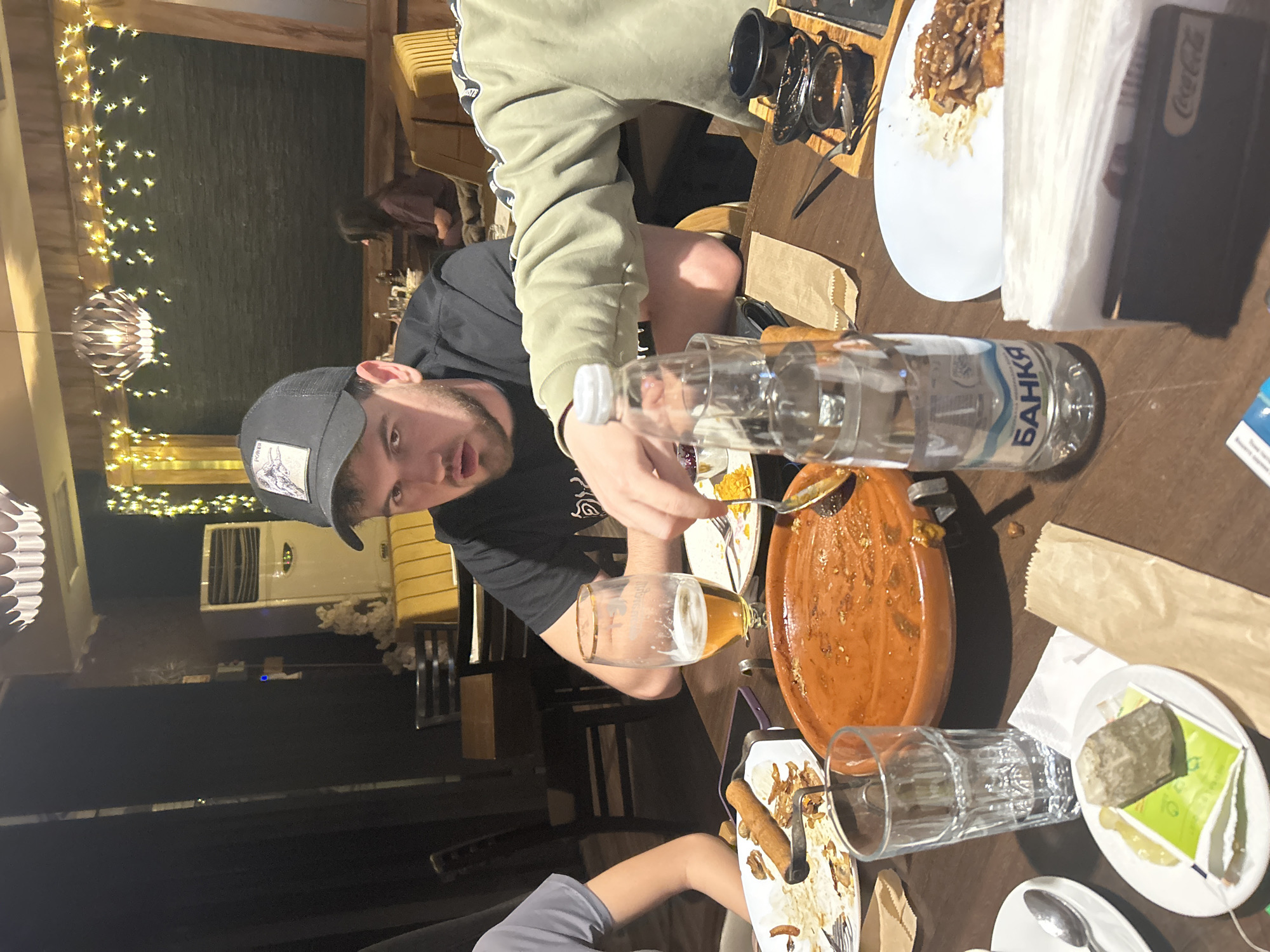 A group of people are dining indoors at a restaurant, possibly in Sofia, Bulgaria. One person is in focus, wearing a black t-shirt and a gray cap, using a spoon to serve themselves from a shared dish. A partially empty glass of beer sits next to them. Other individuals are also at the table. There are plates with leftover food, used napkins, glasses, and a bottle of water on the table. The restaurant is decorated with string lights and has a contemporary design.