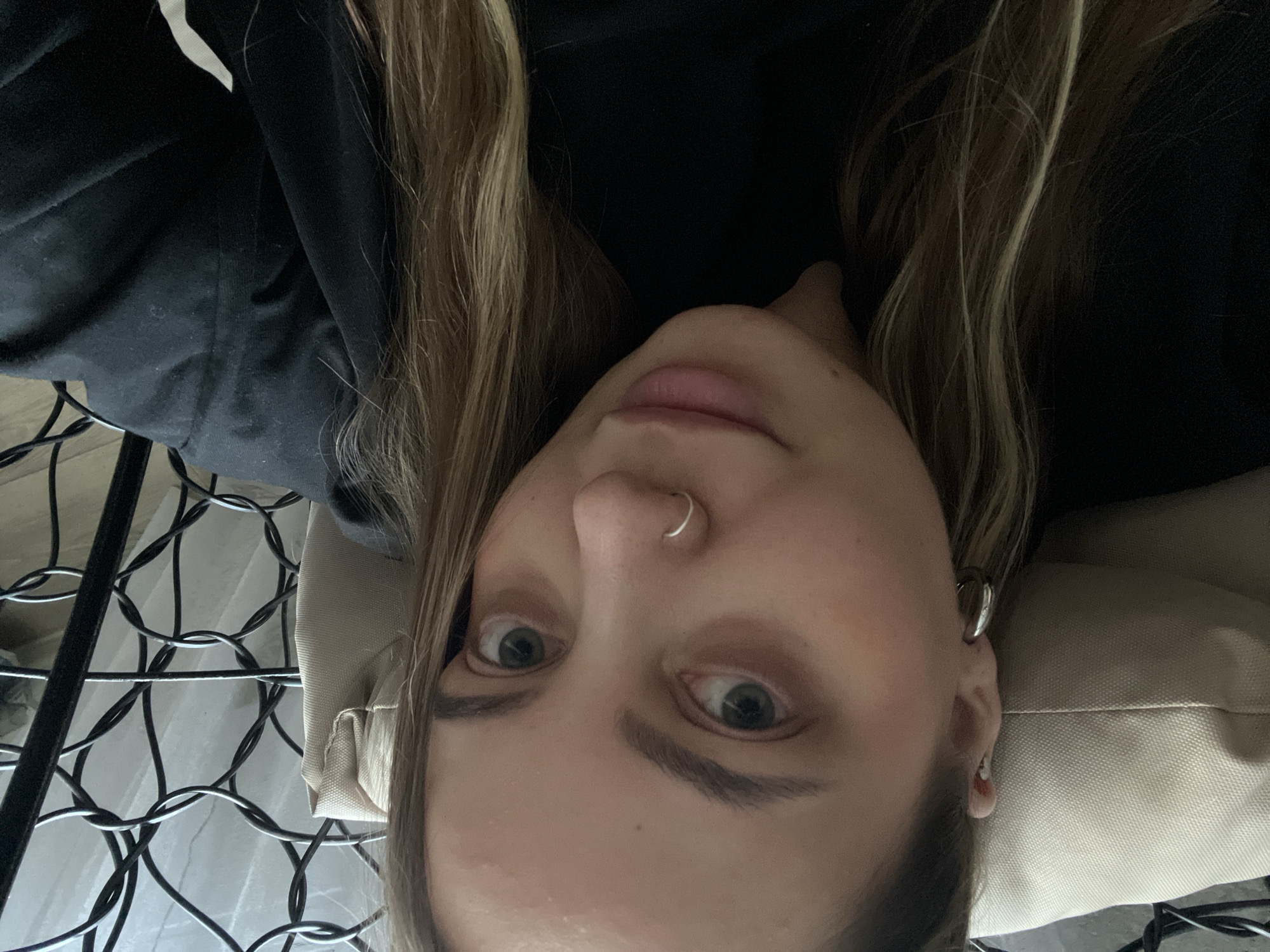 A close-up, selfie-style shot shows a young woman with light brown hair, wearing a black top. She has hoop earrings in both ears and a silver nose ring. Her eyes are looking towards the camera, and her lips are slightly parted. She appears to be sitting in an armchair with a light-colored cushion. The background includes a dark, ornate metal structure, possibly part of the chair, and a white curtain or window covering. The overall lighting suggests an indoor environment. This image does not contain any information that would allow for determination of its location within Novopokrovskaya, Russia.