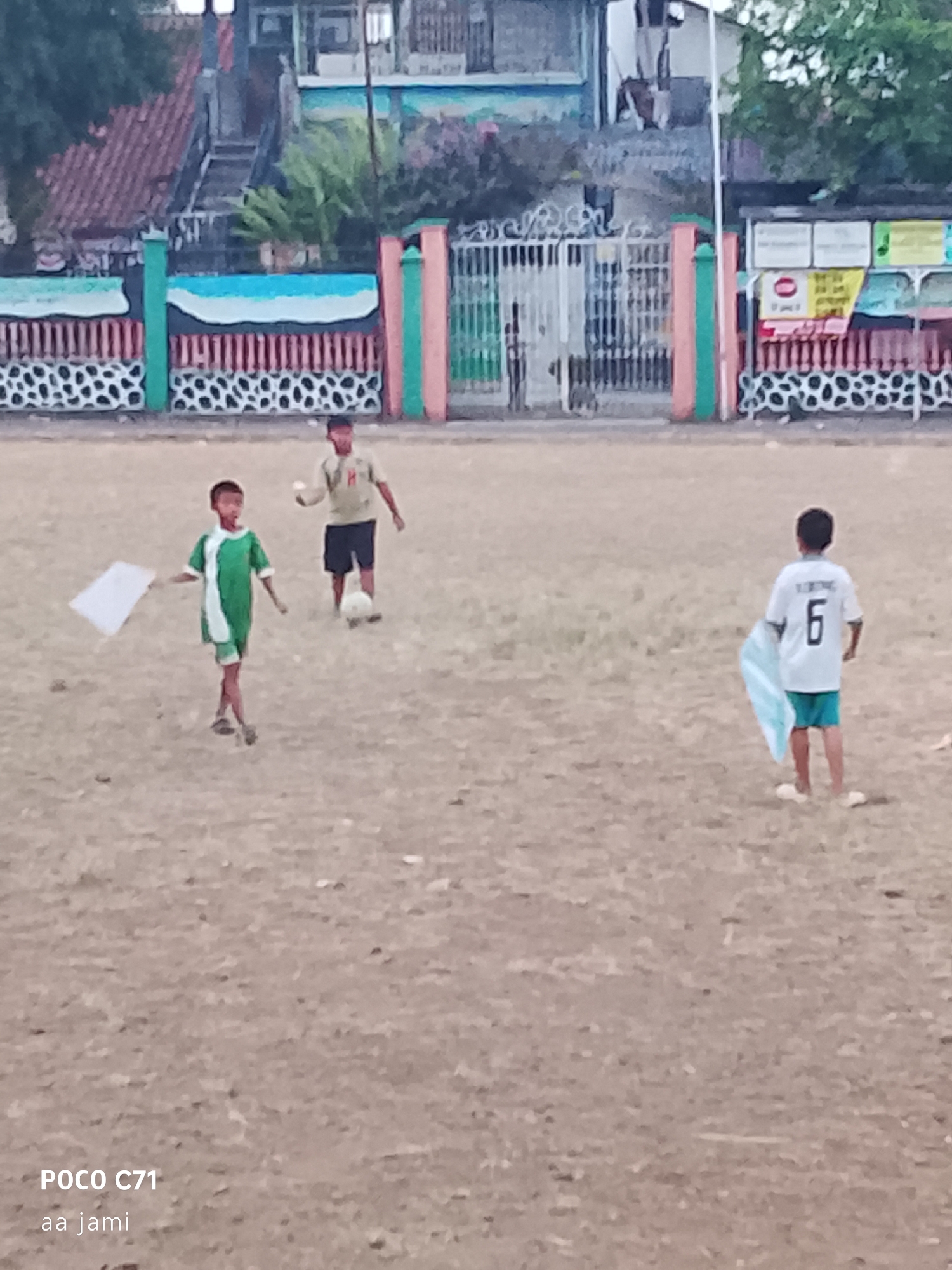 The image depicts an outdoor scene of children playing soccer on a dirt field, likely in Tasikmalaya, Indonesia, given the "POCO C71 aa jami" watermark, which suggests a location in Indonesia. Three young boys are visible. One boy, in the foreground and to the left, wears a green and white soccer jersey and shorts, and holds a white piece of paper. Another boy, in the mid-ground, wears a light-colored shirt and dark shorts, kicking a soccer ball. To the right, a third boy, wearing a white jersey with the number "6" on the back and teal shorts, faces away from the camera. In the background, a fence with colorful decorations and a building are present. The ground is a light brown, dusty dirt field, common for informal soccer games in the region. The overall lighting suggests it may be late afternoon or early evening.