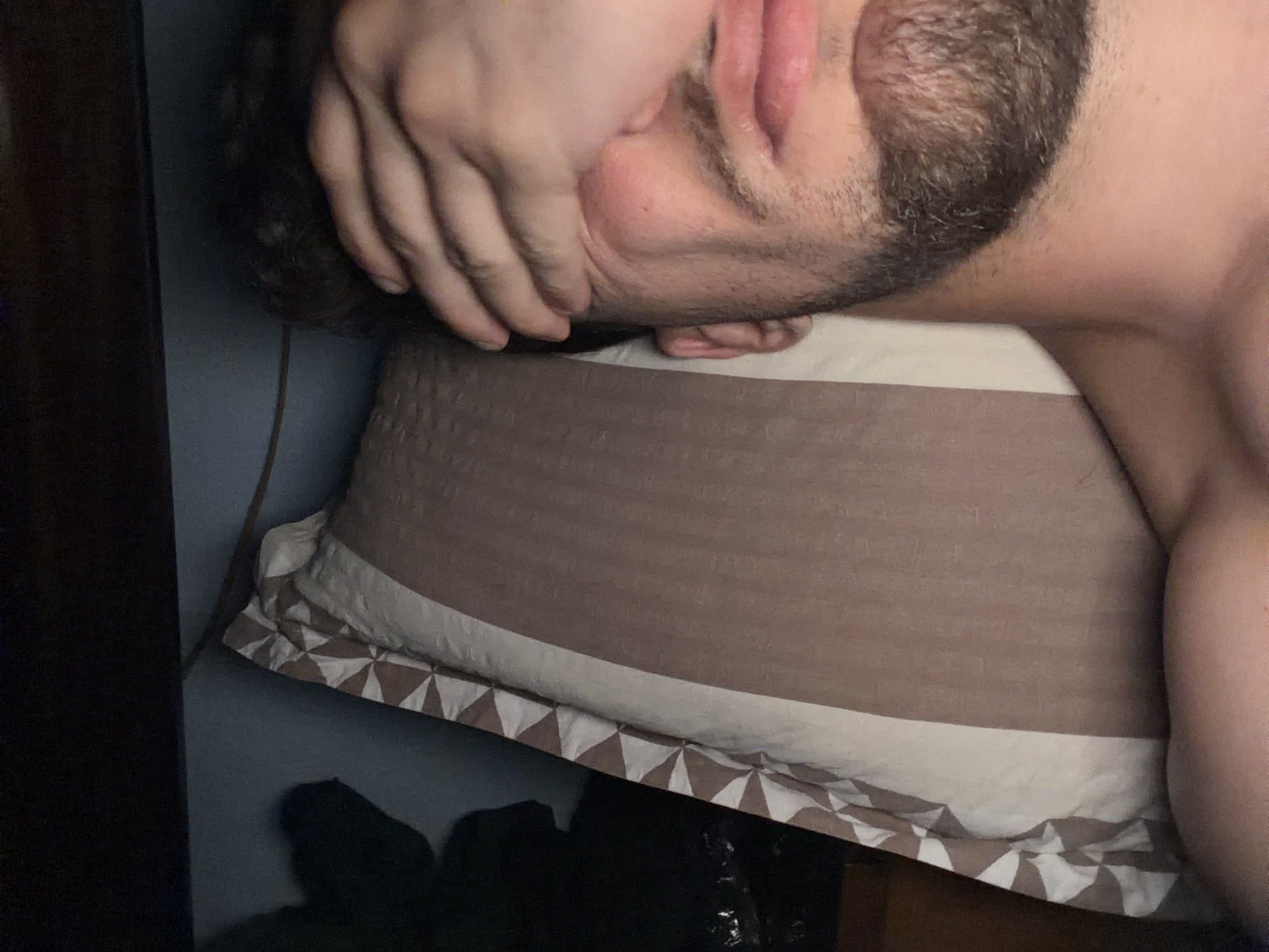 A close-up, overhead shot captures a person lying on a pillow, with their hand covering their eyes. The pillow has a brown and cream striped pattern with a decorative geometric border on one side. The background appears to be a dark wall and a dark wooden surface. The person has a beard and visible skin on their shoulder and upper chest. The scene is dimly lit, suggesting it might be nighttime or a dark room. No specific location in Tsarevo, Bulgaria is indicated in the image.