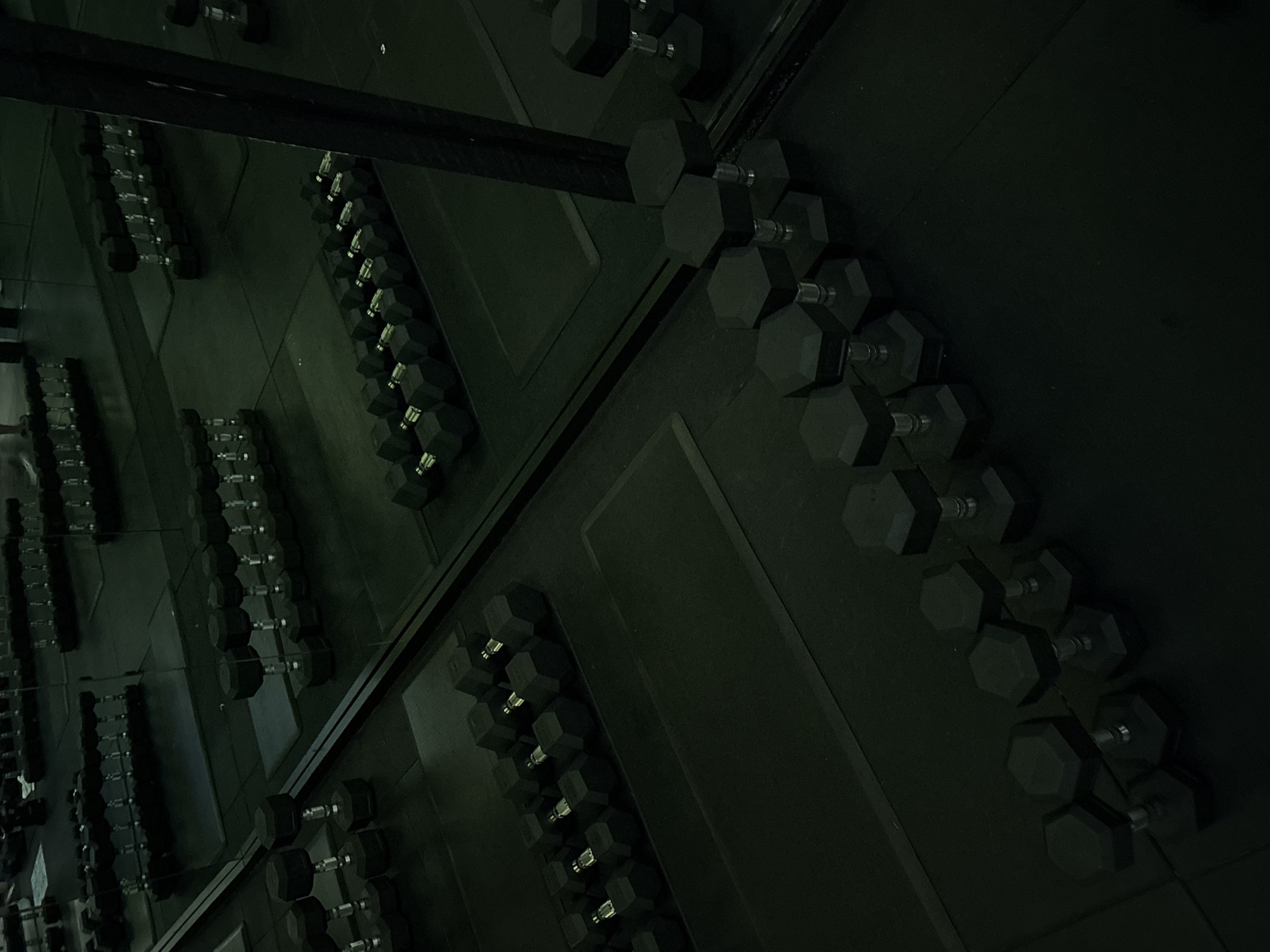 This low-light interior photograph captures a gym or weightlifting area, dominated by rows of black hexagonal dumbbells. The dumbbells, featuring metallic handles and rubberized heads, are neatly arranged on a dark, possibly rubberized, floor. Large mirrors on the left reflect the rows of weights, creating a sense of an expansive space filled with equipment and amplifying the geometric patterns formed by the dumbbells. A dark, horizontal beam or bar extends across the upper section of the image.
The overall color palette is very dark, primarily black and deep greys, contributing to a stark and intense atmosphere. The dim lighting highlights the metallic elements of the dumbbells and the edges of their hexagonal heads, but most of the scene remains in shadow. There are no people visible, suggesting a quiet moment in the gym, devoid of active training. No visible text is present in the image. The scene is set in what is described as San Bernardino Tlaxcalancingo, Mexico, and evokes a feeling of seriousness and readiness for physical exertion.