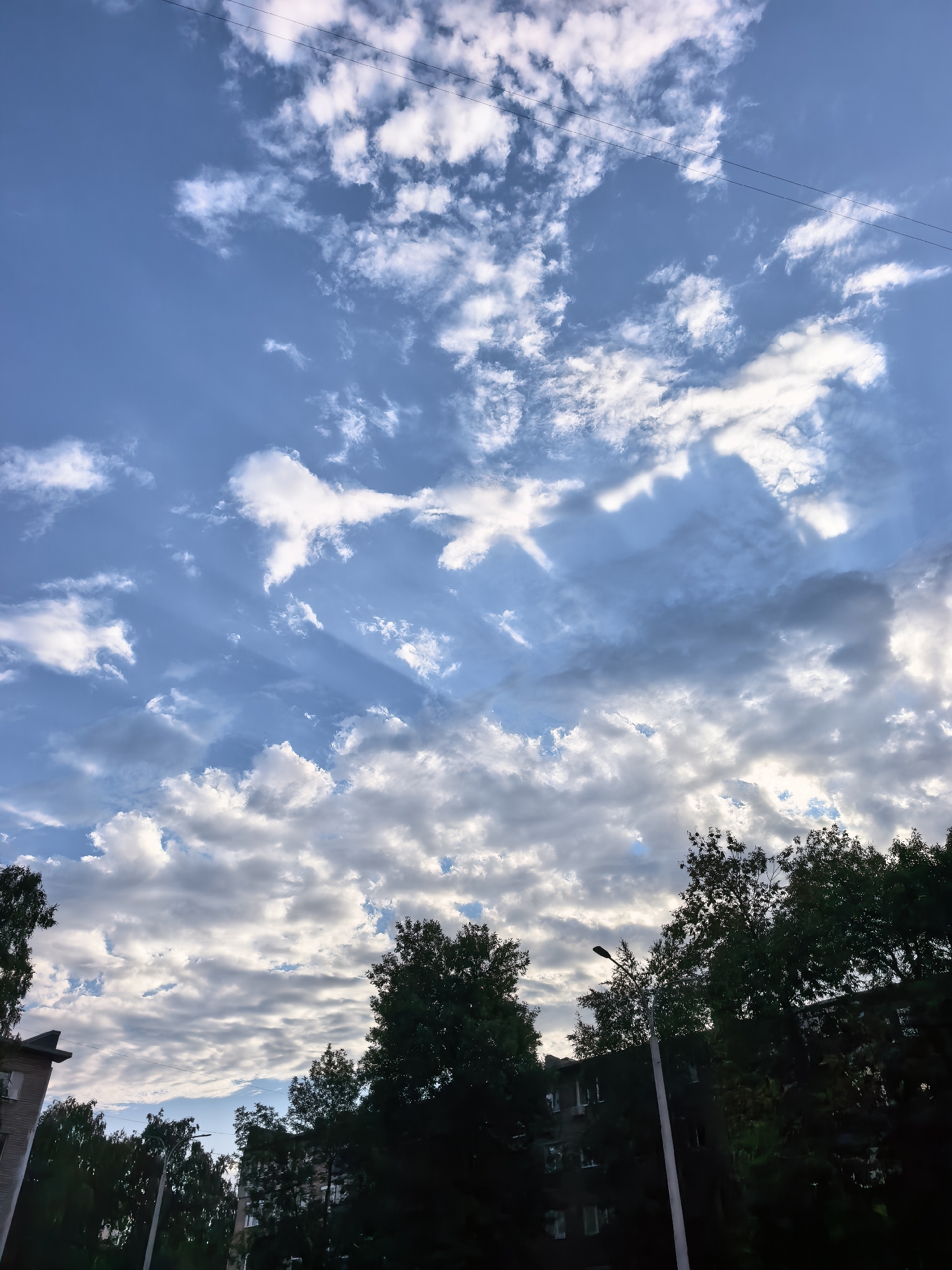 The image captures a dramatic sky over Sterlitamak, Russia, with a mix of bright white and dark grey clouds against a blue sky. Sunbeams pierce through the clouds, creating a striking visual effect. Silhouetted against the sky are the tops of lush green trees and the dark facades of apartment buildings. A single streetlamp is visible, its pole extending upwards. Power lines are also faintly visible against the sky. The scene suggests an urban environment, possibly a residential area, under a dynamic sky, likely during late afternoon or early evening. There are no people or animals visible in the image.