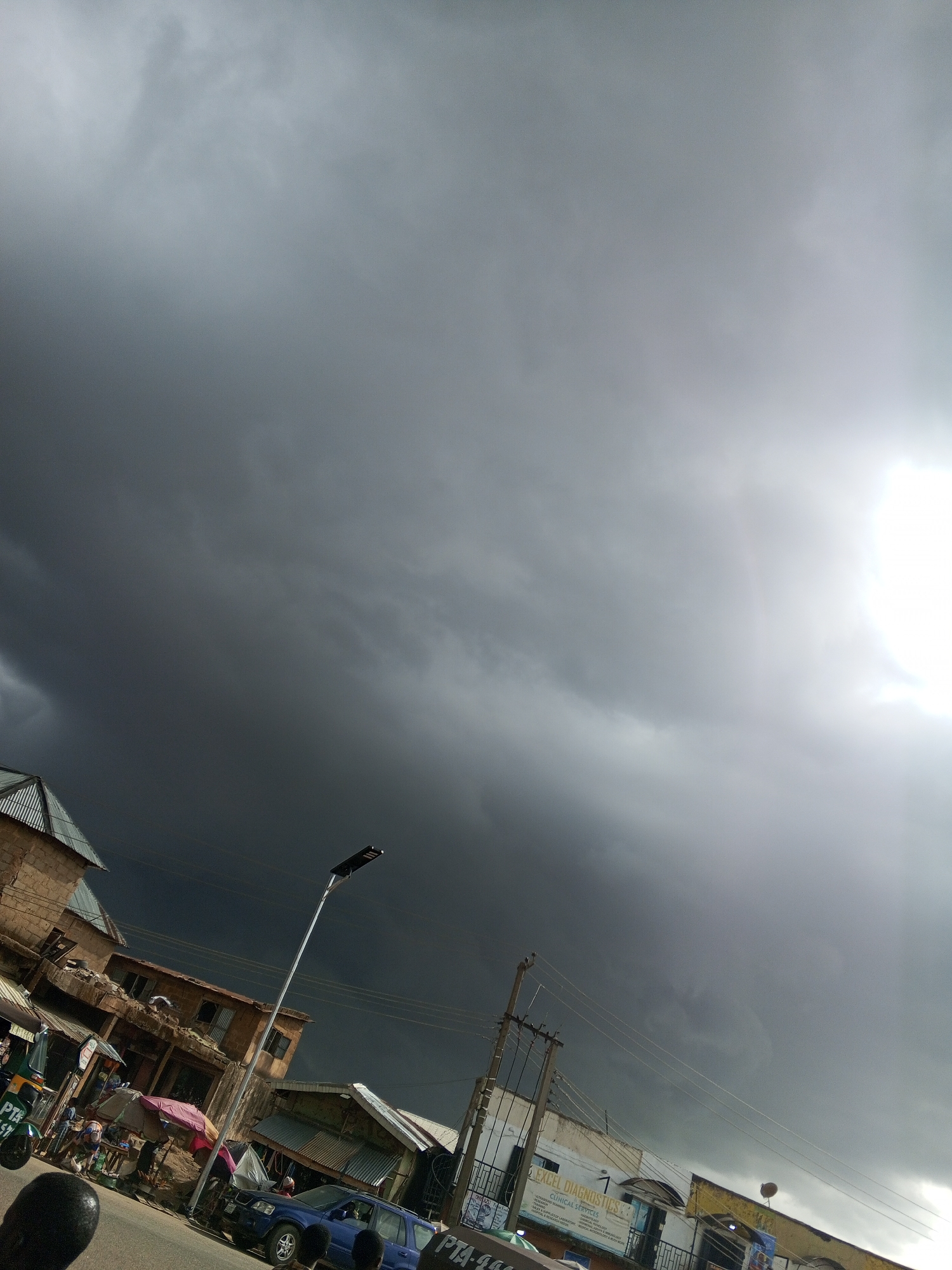 The image captures a street view in Gwong, Nigeria, under a dramatically overcast sky with dark storm clouds dominating the scene. The sunlight is partially obscured, yet it casts a bright haze towards the upper right.
The foreground shows a street scene with buildings on both sides. Structures vary in style, including traditional brick constructions with corrugated metal roofs. Several commercial establishments are visible along the street level.
A few cars and motorcycles are parked on the side of the street. Some people are milling about, including those gathered near street vendors and shops, indicating commercial activity. Utility poles with electrical wires are present, adding to the urban environment. A street light stands tall on the street side.