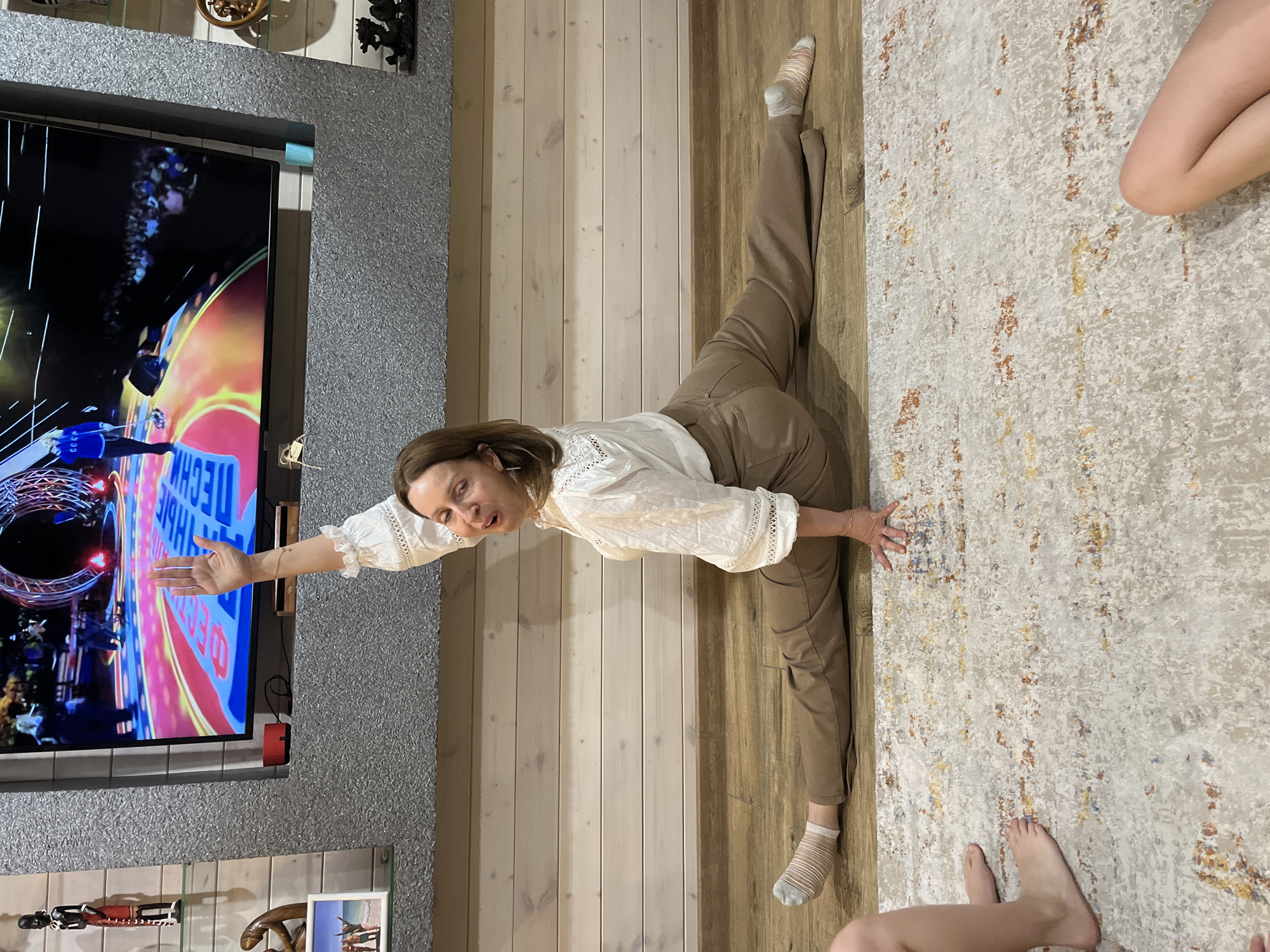 A woman is doing the splits in a room. She is wearing a white blouse and brown pants, with socks on one foot. She is reaching out with her left hand. A television is to her left, displaying a scene with text in Cyrillic script. There is a rug to her right. The room has wood paneling on the floor and wall. There is a shelf below the television with figurines. There are parts of other individuals visible in the rug area. This is unlikely to be in Novaya Vozhoika, Russia, because that is a fictional location.