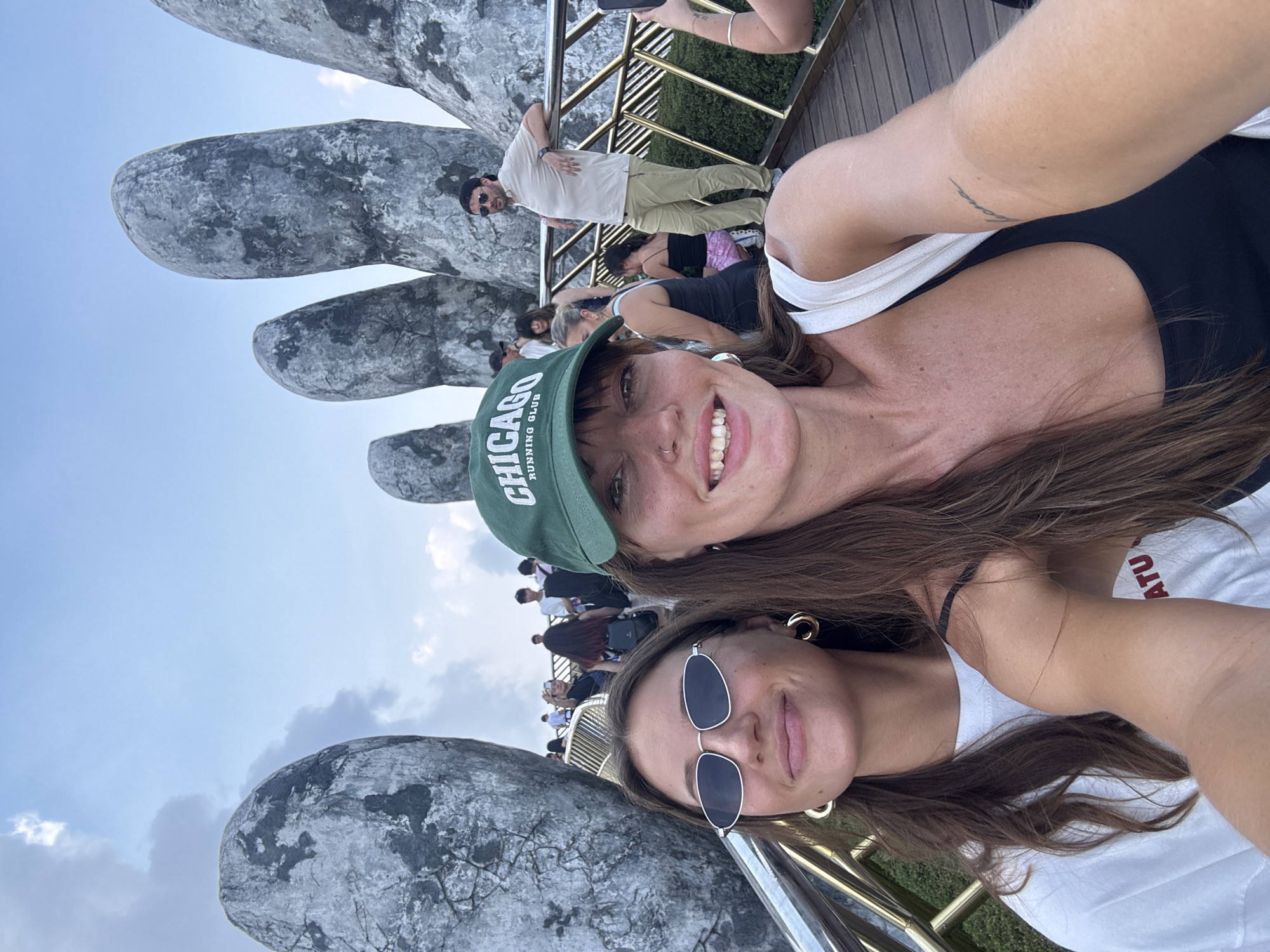 Two women are in a selfie at the Golden Bridge in Đà Nẵng, Vietnam. The bridge is supported by two massive stone hands. The woman on top wears a "Chicago Running Club" baseball cap. The woman below wears dark sunglasses. People populate the bridge in the background.