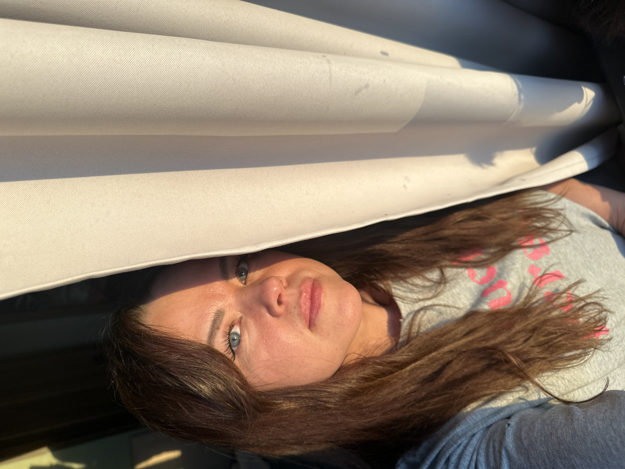 A woman with long, brown hair and striking blue eyes is depicted resting beneath a rolled-up, light-colored fabric structure, possibly an awning or tent. Her head is tilted back, and she gazes directly at the camera with a neutral, serene expression. Warm, golden sunlight illuminates her face, indicating a sunny late afternoon or early morning.
She is wearing a light gray t-shirt with pink lettering that partially spells out "SHORTS" across the chest. Another piece of dark fabric, possibly clothing or a blanket, is visible in the lower right corner of the frame. The fabric above her head exhibits a subtle diagonal weave pattern and shows some faint marks or stains.
The scene suggests an outdoor or semi-outdoor leisure activity, likely in Chaikovsky, Russia, where the woman is relaxed and enjoying the warm weather.