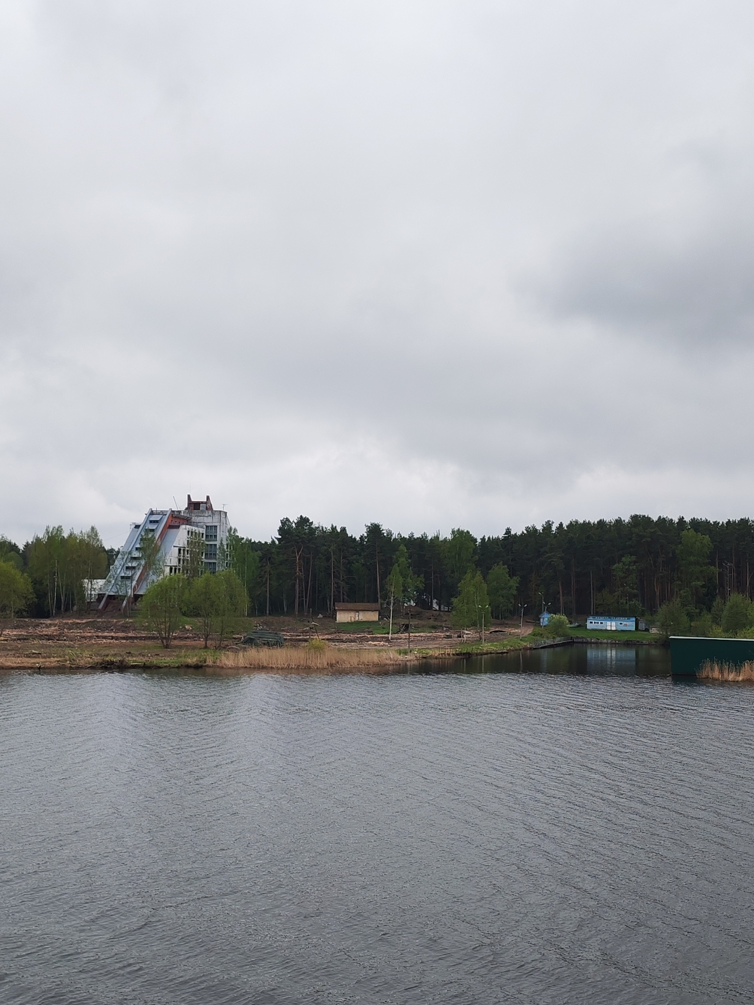 The image shows a large body of water under an overcast sky. A building with a distinctive step-like structure is visible on the left, with a dense forest behind it. Other small structures and vegetation are scattered along the shoreline. The location is Novomelkovo, Russia.