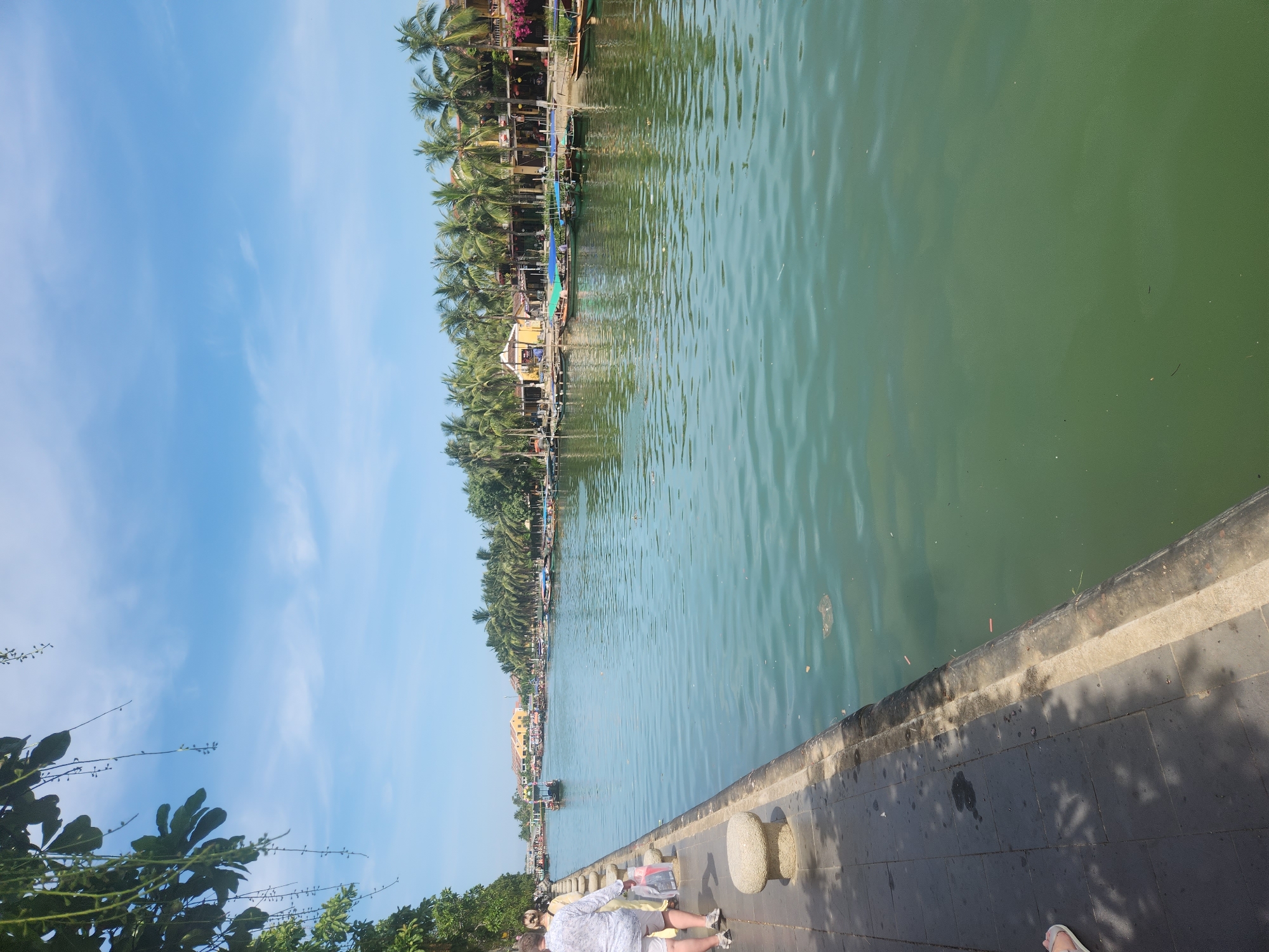 The image shows a wide, calm canal in Hội An Ward, Vietnam, with boats docked along the opposite bank. A paved walkway runs along the near side of the canal, with people walking and standing. Palm trees line the buildings on the far bank, casting shadows on the water. The water is a greenish-blue color and reflects the sky. The sky is bright blue with scattered white clouds. A small boat is in the middle of the canal, heading away from the viewer. The overall scene is one of a peaceful riverside setting.