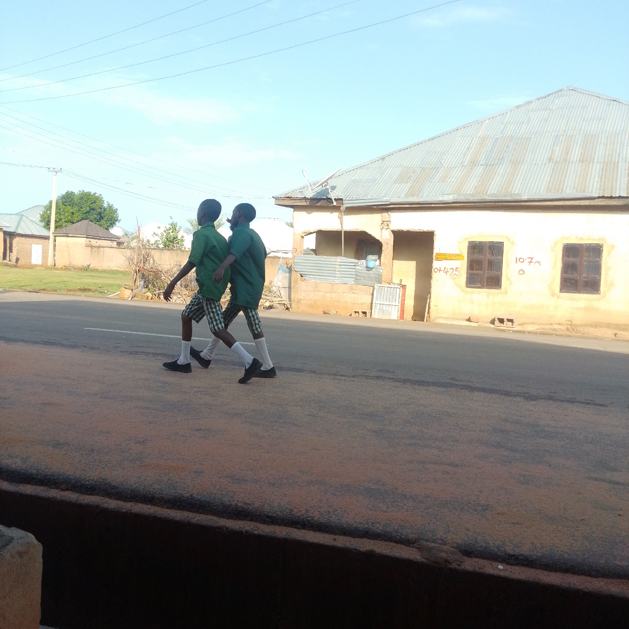 Two young boys, dressed in green school uniforms with checkered shorts and white socks, walk side-by-side on a paved road. The boy on the left wears a short-sleeved green shirt and black shoes, while the boy on the right has a longer-sleeved green shirt and black shoes. They are walking from left to right in the frame. Behind them, a low-rise building with a corrugated metal roof is visible, painted yellow with visible signs of weathering and some brickwork exposed. There are two dark-framed windows on the facade of this building. Graffiti, including "10.7M" and what appears to be "ALUA SAYA KUSA J +425", is present on the building's wall. The environment is outdoors, under a clear blue sky with some power lines strung across the top of the image. The road surface is asphalt, with a white line painted down the center. To the left of the building and further in the background, other structures and trees are visible, suggesting a semi-urban or rural setting. The foreground shows a darker, textured surface, possibly a curb or raised planter box. This scene is located in Gombe, Nigeria.