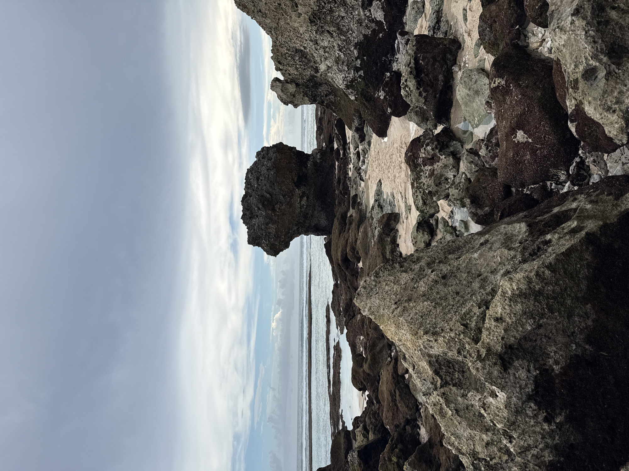 The image depicts a coastal scene in Ungasan, Indonesia. Large, dark grey rocks and boulders dominate the foreground and right side of the frame. In the center, a distinct, mushroom-shaped rock formation stands prominently against the sky and ocean. The ocean is visible as a narrow horizontal strip between the rocks. The sky is overcast with grey and white clouds. No people are visible.