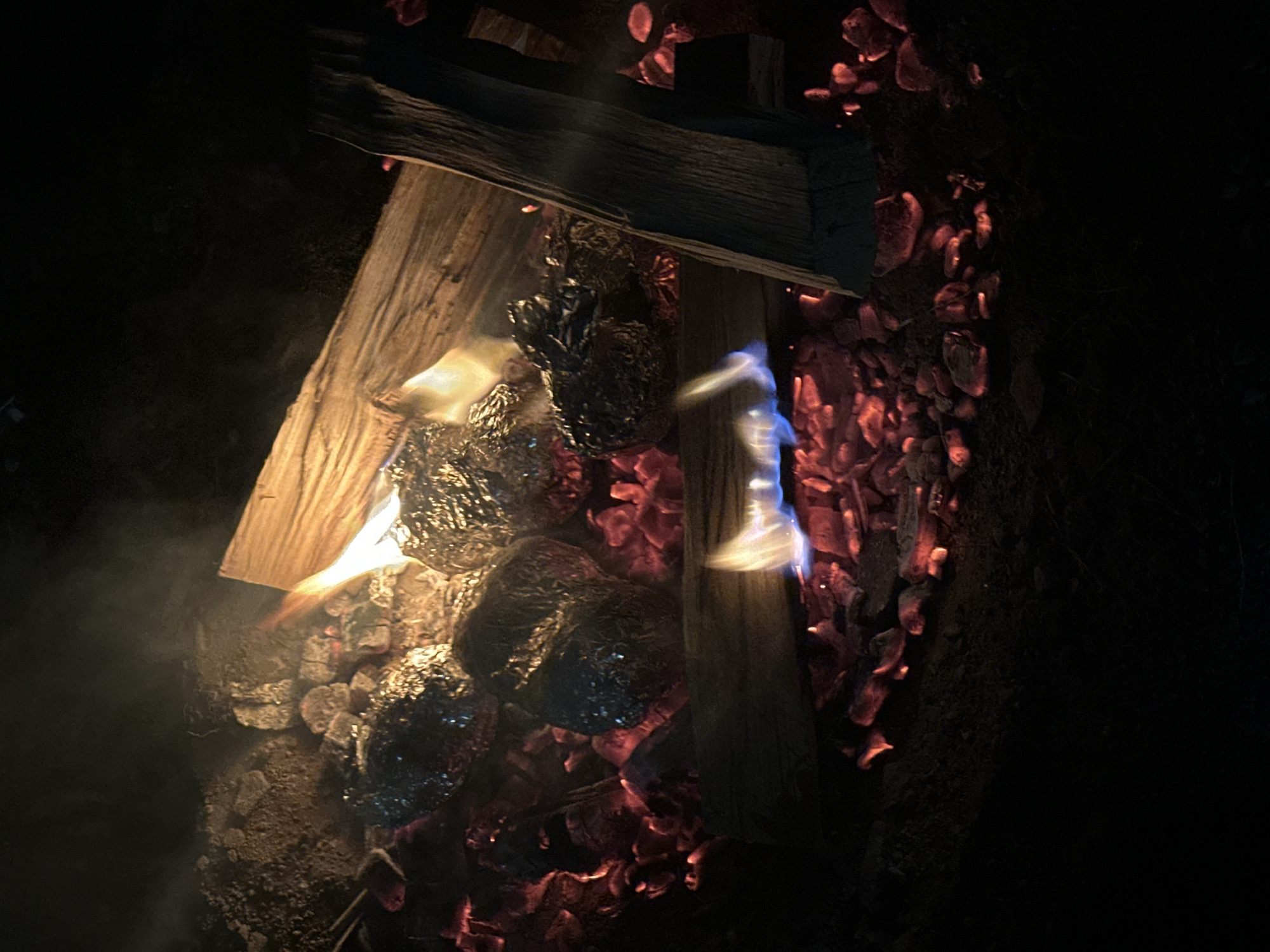 The image captures a close-up view of potatoes wrapped in aluminum foil being cooked in a campfire. The fire is actively burning, with glowing red embers and visible flames. Several logs are present, some charred and others appearing freshly placed. The potatoes are nestled amongst the hot coals, indicating they are in the process of being baked. The surrounding environment appears to be dark, likely night, with the light from the fire illuminating the immediate area. There is no indication of the specific location within the city of Жемчужный, Russia, based solely on the visual information provided. The scene is purely focused on the cooking process.