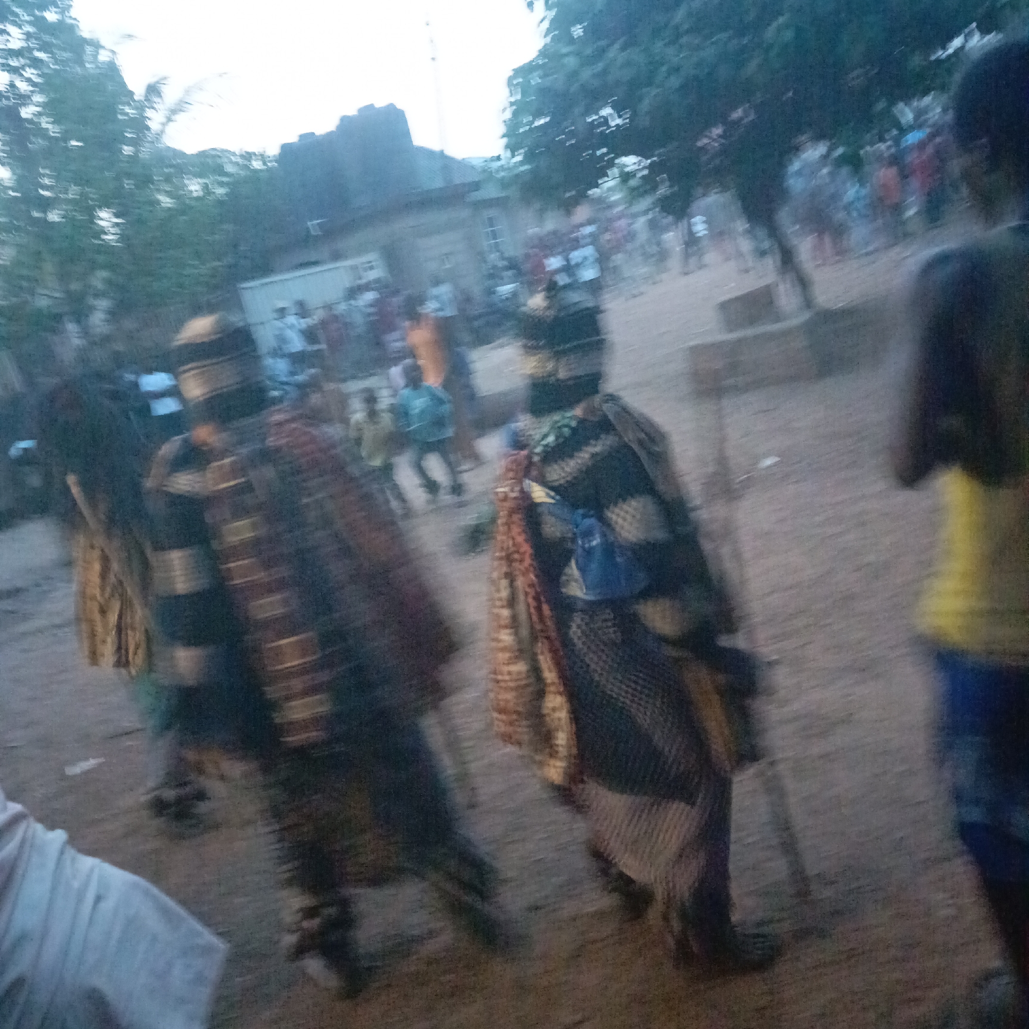 The image shows several individuals dressed in traditional attire in what appears to be a public space. People in the background seem to be observers. Based on the prompt, this is likely a cultural event in Akwanga, Nigeria. The overall environment seems to be a dusty or sandy area, possibly a village square or open field. The lighting suggests it might be late afternoon or early evening. The image quality is somewhat blurry, possibly due to movement or camera shake.