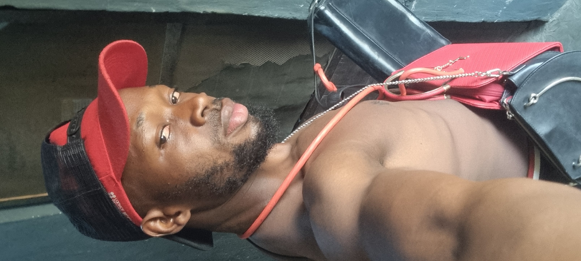The image shows a dark-skinned man wearing a red and black baseball cap. He has a beard and is topless. Straps of what appears to be two bags are across his chest and shoulder. One bag is red and the other is black. The background is indistinct, with a greyish wall. There is no specific evidence in the image to confirm its location within Ikeja, Nigeria.
