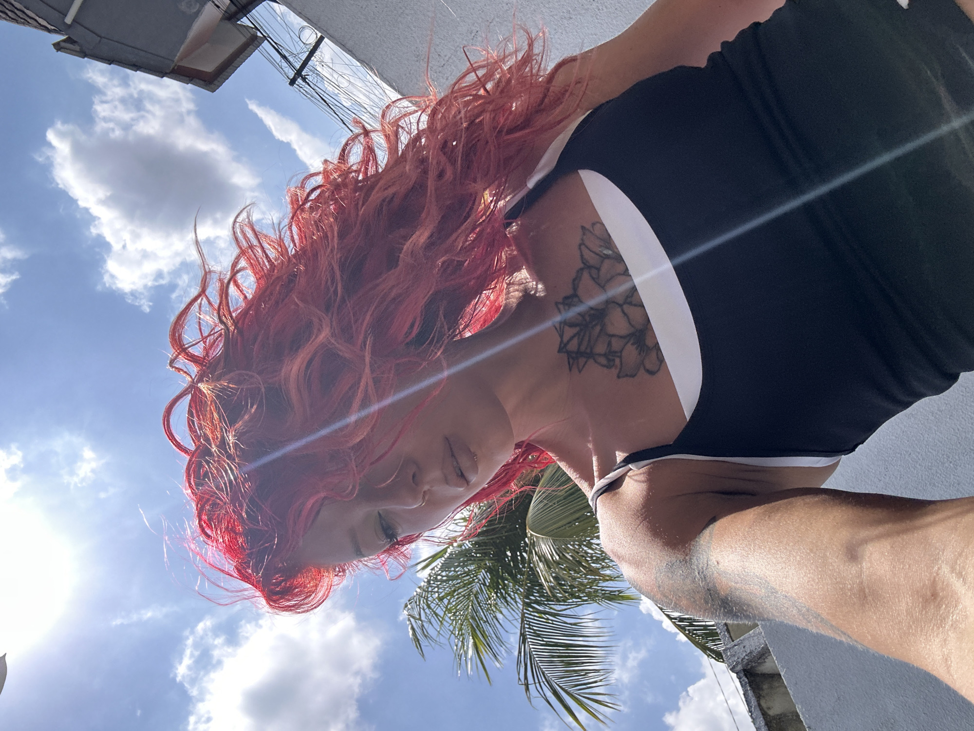 A medium shot captures a woman with vibrant red, wavy hair against a bright blue sky dotted with white clouds and the sun. She is looking downwards, with her head tilted. The woman is wearing a black tank top with a white band at the top, and a white undergarment. She has a tattoo on her chest and another on her upper arm. To the left of the woman, a palm tree is visible. A white wall runs along the bottom and right side of the frame, and a building with a gray roof is partially visible in the upper right. Electrical wires are strung across the background. The scene appears to be outdoors in a sunny, urban or suburban environment, potentially in the city of North Kuta, Indonesia, given the presence of palm trees and architecture.