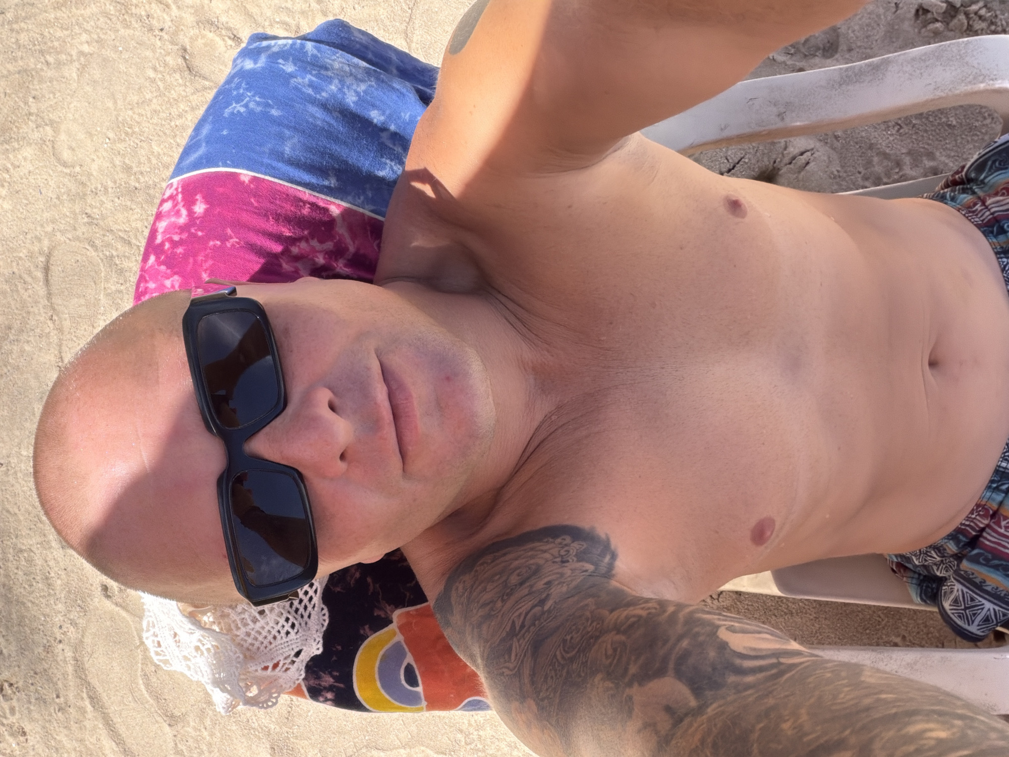 A man with a shaved head and sunglasses is lying on a beach chair on a sandy beach. He is shirtless and wearing patterned swim trunks. A colorful towel is behind his head. The location is Choeng Thale, Thailand.
