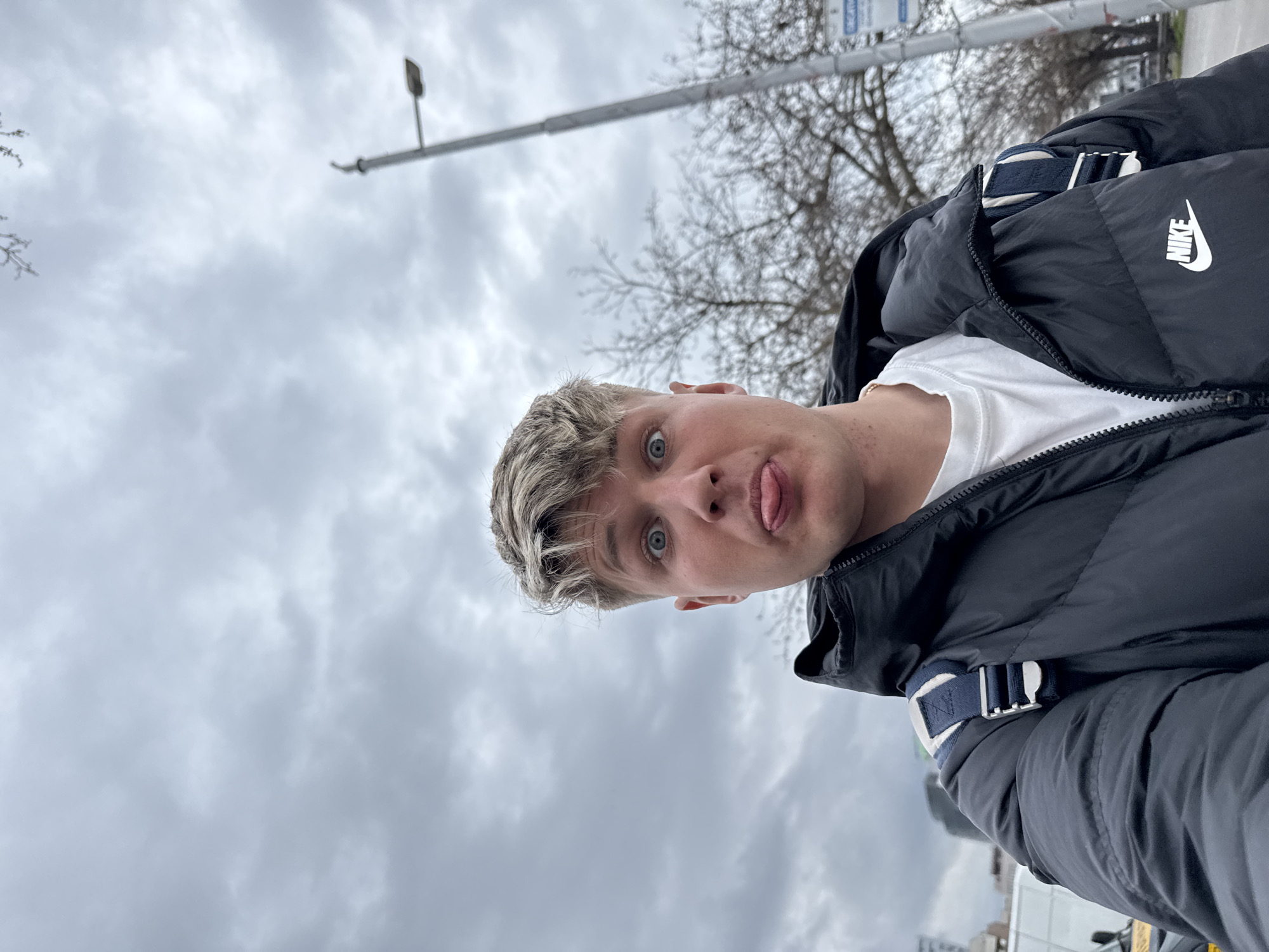 An upward-angled, eye-level selfie-style photograph captures a young adult male on a street in Plovdiv, Bulgaria, beneath an overcast sky. The subject, positioned centrally, has fair skin, light blonde hair with a textured fringe, and wide blue eyes. He wears a black puffer jacket with a white Nike logo on the left chest and a white crew-neck t-shirt. A blue and white backpack strap is visible over his right shoulder. His expression includes wide eyes and his tongue extended, indicative of a playful or humorous gesture.
The background consists of a blurred urban environment. To the subject's left, a light-colored vehicle, possibly a white van, and indistinct building structures are visible. To his right, a grey street light pole with a single luminaire extends into the frame. Bare tree branches, suggesting a dormant season, are scattered across the midground. A blue rectangular sign partially visible in the lower right background reads "GREAT HOTEL" in white capital letters, with Cyrillic text below, indicating the Grand Hotel Plovdiv. The sky is entirely covered with thick, grey clouds.