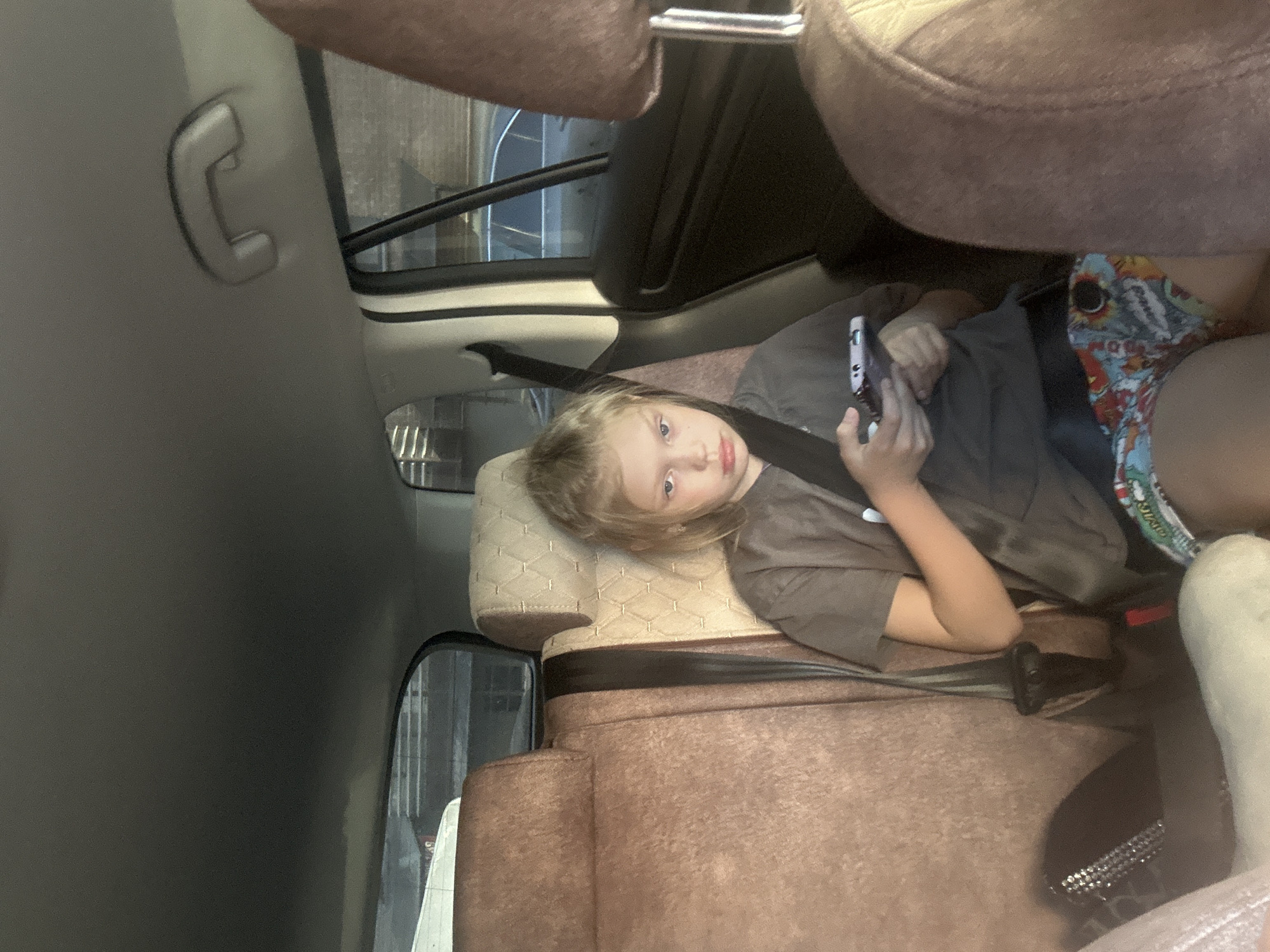 A young girl with blonde hair is seated in the back of a car, wearing a grey t-shirt and patterned shorts. She is holding a smartphone in her hands and looking at it. The interior of the car has tan and brown upholstery. The girl is wearing a seatbelt. The background outside the car windows shows buildings and other vehicles, suggesting an urban environment. This scene is likely taking place in the city of Abakan, Russia.