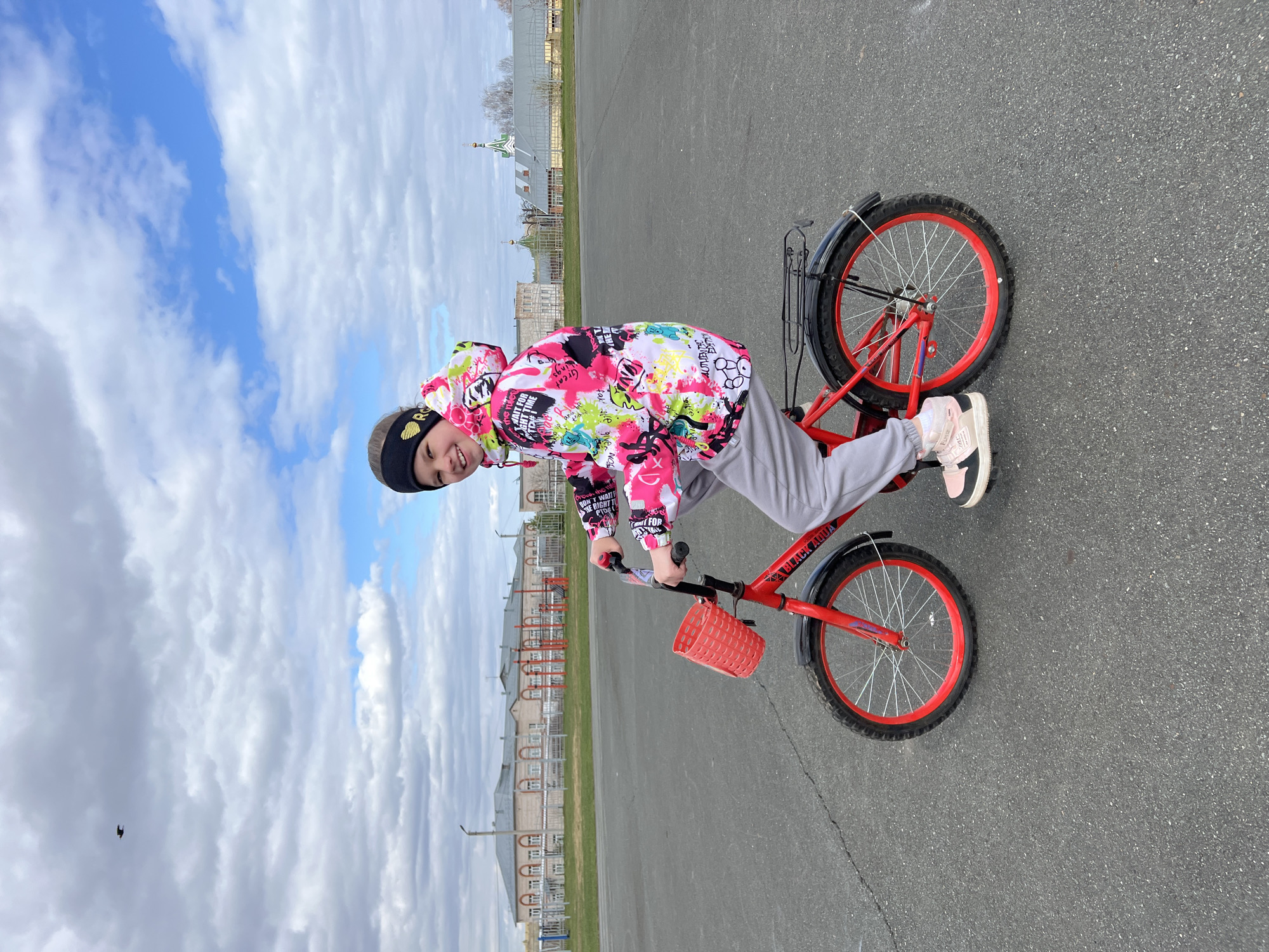 A medium shot shows a girl riding a red bicycle in Перевозное, Russia. The girl is wearing a black headband, a colorful jacket, and gray pants. The red bicycle has a basket in the front. The environment appears to be a flat asphalt area with buildings visible in the background. The sky above is partly cloudy.
