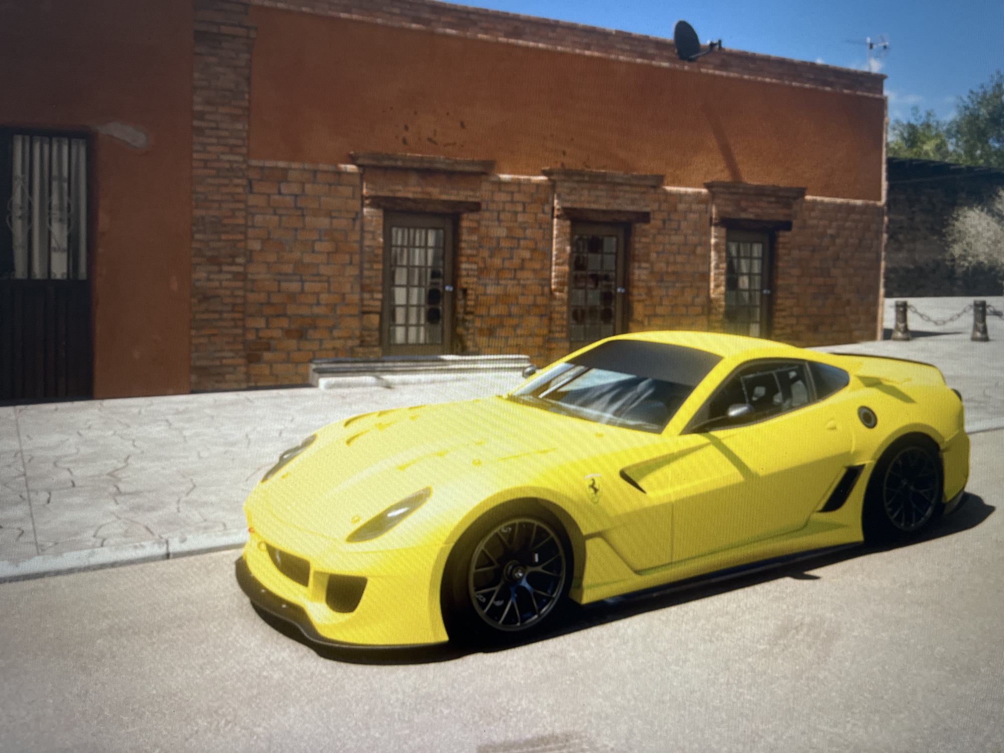 The image shows a bright yellow Ferrari parked on a street in front of a two-story building. The building has an orange-colored facade with a brick lower section. There are three dark-colored doors on the lower level, each with a grid pattern in the glass. The street is paved with light-colored cobblestones and asphalt. In the background, trees and a bright blue sky are visible. The car is a sports car with a low profile and sleek design. There are no people visible in the image. The scene appears to be a sunny day.