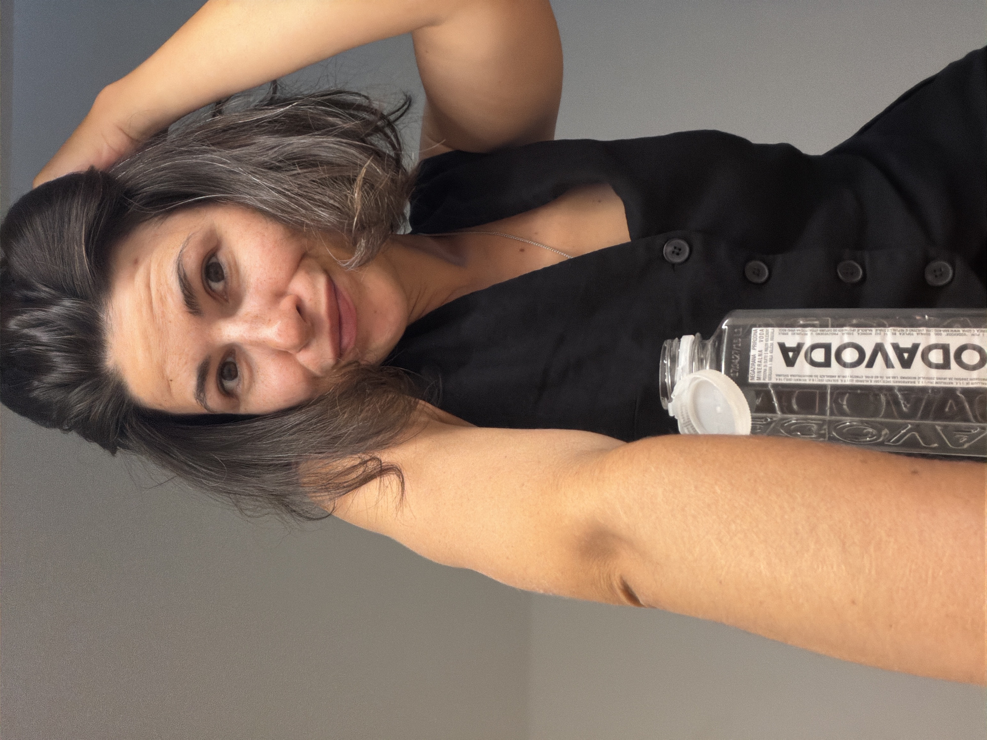 This portrait-oriented selfie features a woman indoors, looking directly at the camera with a gentle, friendly smile. She has dark, wavy hair with noticeable streaks of gray, and her brown eyes are prominent. She is wearing a dark, possibly black, sleeveless top or dress with buttons down the front, and a thin silver chain necklace. Her left arm is raised above her head, while her right arm is positioned alongside a clear plastic water bottle with a white cap.
The background is a plain, light gray wall. The lighting is soft and even, suggesting an indoor setting. The label on the water bottle is visible but appears reversed due to the selfie camera. The prominent brand name visible is "ODAVODA" (appearing as ADAVODO). Other words clearly discernible on the label, also reversed, include "NEGAZIRANA," "PRIRODNA," "MINERALNA," "VODA," and "IZVORNA," indicating it is natural, non-carbonated mineral spring water. A key detail on the label is "ODAVODA d.o.o. BUDVA" and "CRNA GORA," directly linking the product and its origin to Budva, Montenegro, consistent with the provided location context. The woman's expression conveys a sense of calm and relaxation in this casual moment.