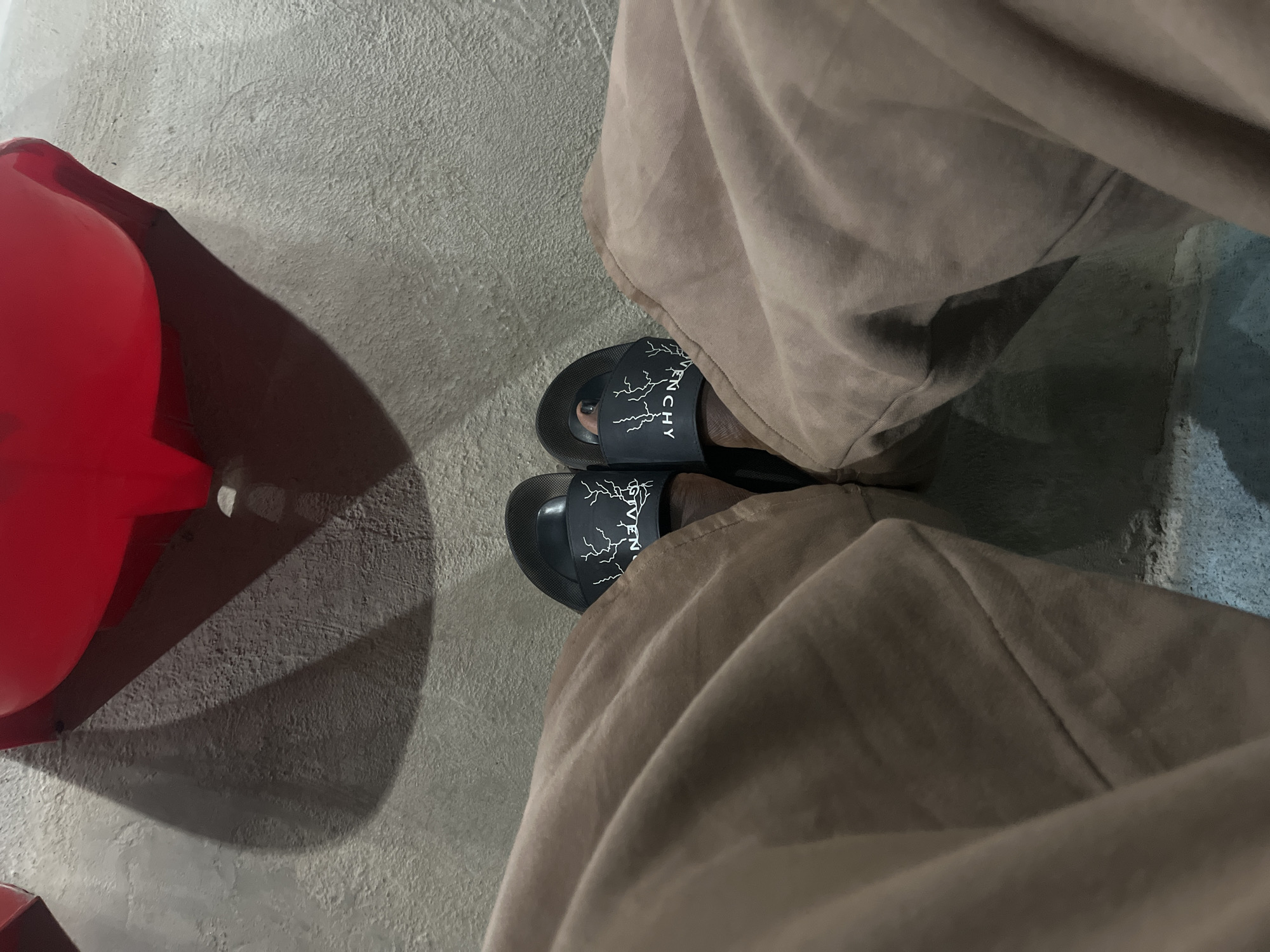 From an elevated perspective, the image captures a person seated on what appears to be a red chair, partially visible on the left. The individual is wearing loose-fitting, brown-beige trousers, the fabric rumpling around their legs. The person's feet are adorned with black sandals featuring a white lightning bolt design and "GIVENCHY" imprinted on the upper portion. The sandals are resting on a gray concrete floor, which extends into the background. The environment is dimly lit. The location appears to be urban, though the specific city and neighborhood are not identifiable from the provided visual information.