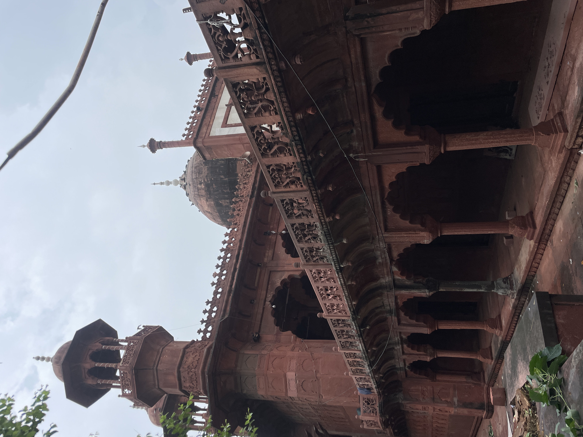 The image shows a close-up view of a red sandstone building in Agra, India, with an overcast sky above. The architecture features intricate carvings and decorative elements typical of Mughal style. On the left, a tall octagonal tower with a domed roof and ornate railings rises above the main structure. The main building has a prominent dome, flanked by smaller finials. A covered walkway with arched colonnades runs along the base of the building, supported by carved pillars. An elevated balcony with decorative railings overlooks the walkway. The ground in front of the building is paved with red stone and shows signs of some debris, including leaves and scattered bricks. The overall impression is one of historical grandeur and architectural detail.
