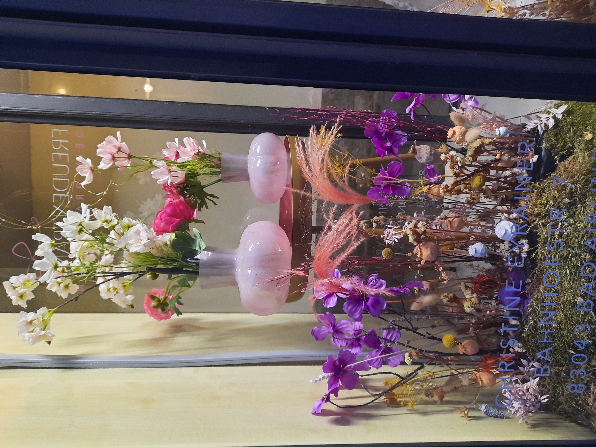 The image shows a shop window display. The display features several vases of flowers, including pink and white flowers in two light pink vases at the top. Below these, there is an arrangement of dried flowers and grasses in shades of purple, pink, and brown. Text is visible on the glass of the window, reading "CHRISTINE EYR BAHNOFSTR 7 83043 BAD A." This text indicates the shop's name, street, and postal code, suggesting the location is Christine Eyr, Bahnhofstraße 7, 83043 Bad Aibling, Germany. The display is set against a yellow wall on the left and a dark blue curtain on the right.