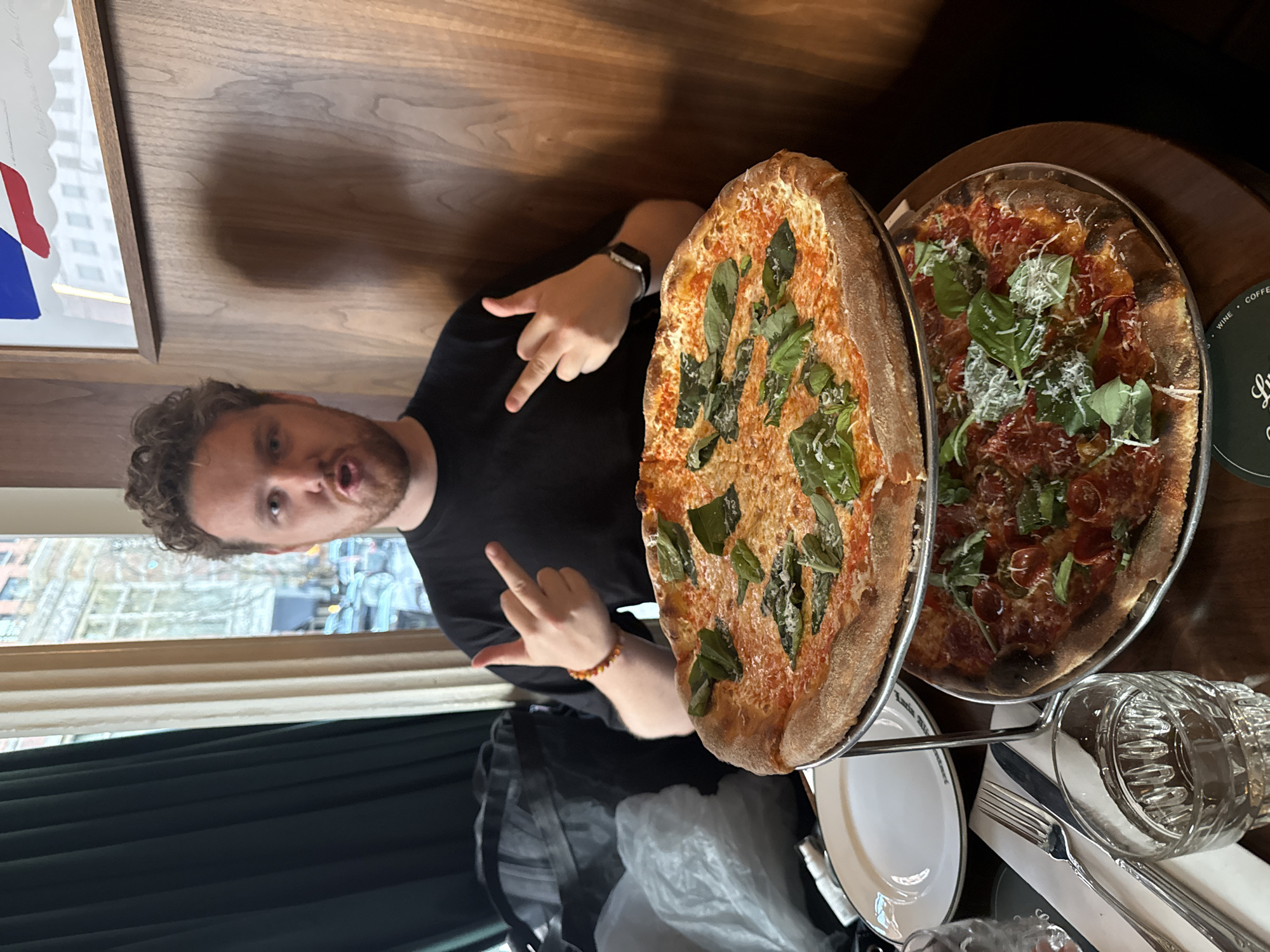 A man with curly light brown hair and a black t-shirt, wearing a watch and a bracelet, is making a hand gesture towards the camera in what appears to be a restaurant. He is situated in front of a tiered stand holding two pizzas. The pizza on the upper tier is topped with mozzarella and basil, while the pizza on the lower tier includes pepperoni, basil, and parmesan cheese. Below the pizza stand, there is a white plate, a wrapped silverware set, and a glass of water on a dark wooden table. Through the window behind the man, blurred buildings and street scenes are visible, suggesting an urban environment. There is also a painting on the left side of the picture. A partial view of a window and an interior curtain can be seen. Based on available information, the location is likely New York City, United States of America.